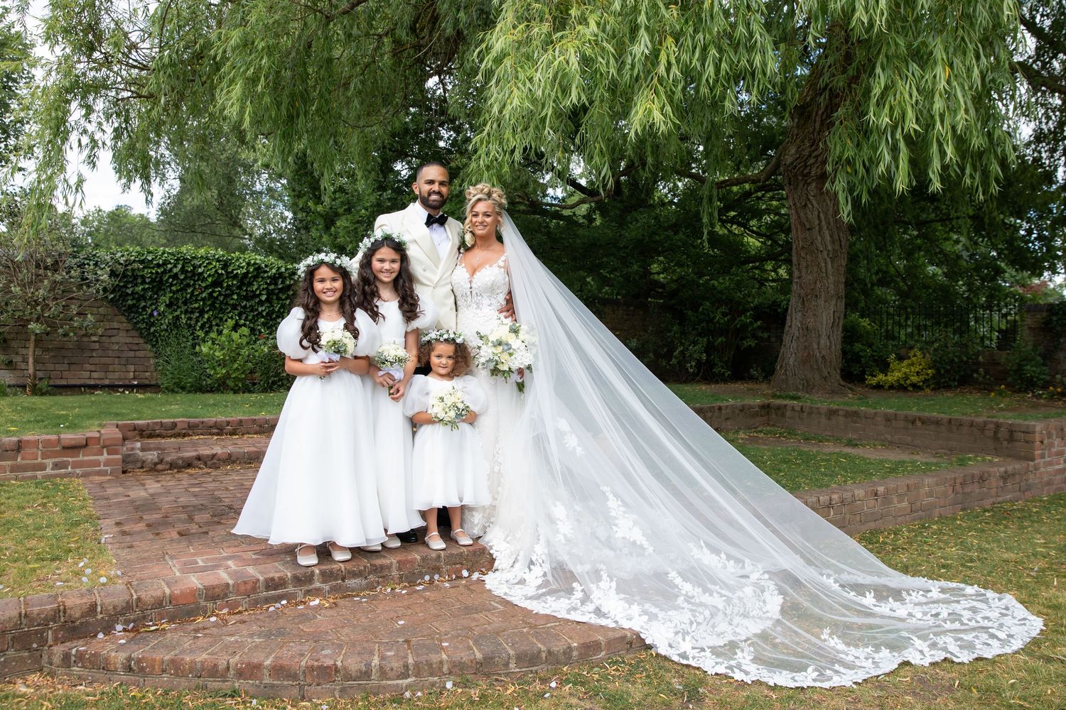 A wedding party poses in a lush garden setting with a flowing white cathedral veil trailing behind them.