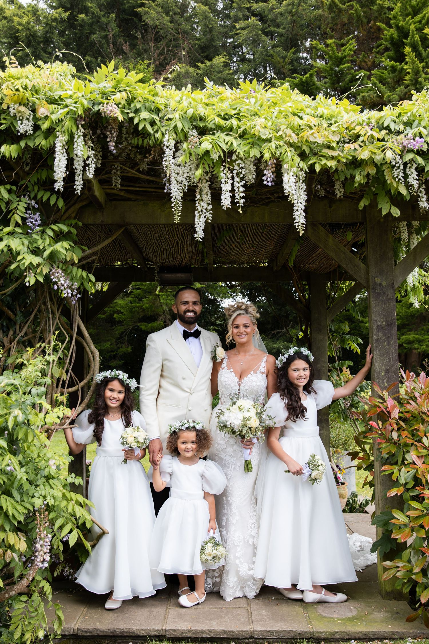 A wedding party poses under a beautiful wisteria-covered pergola in white formal attire.