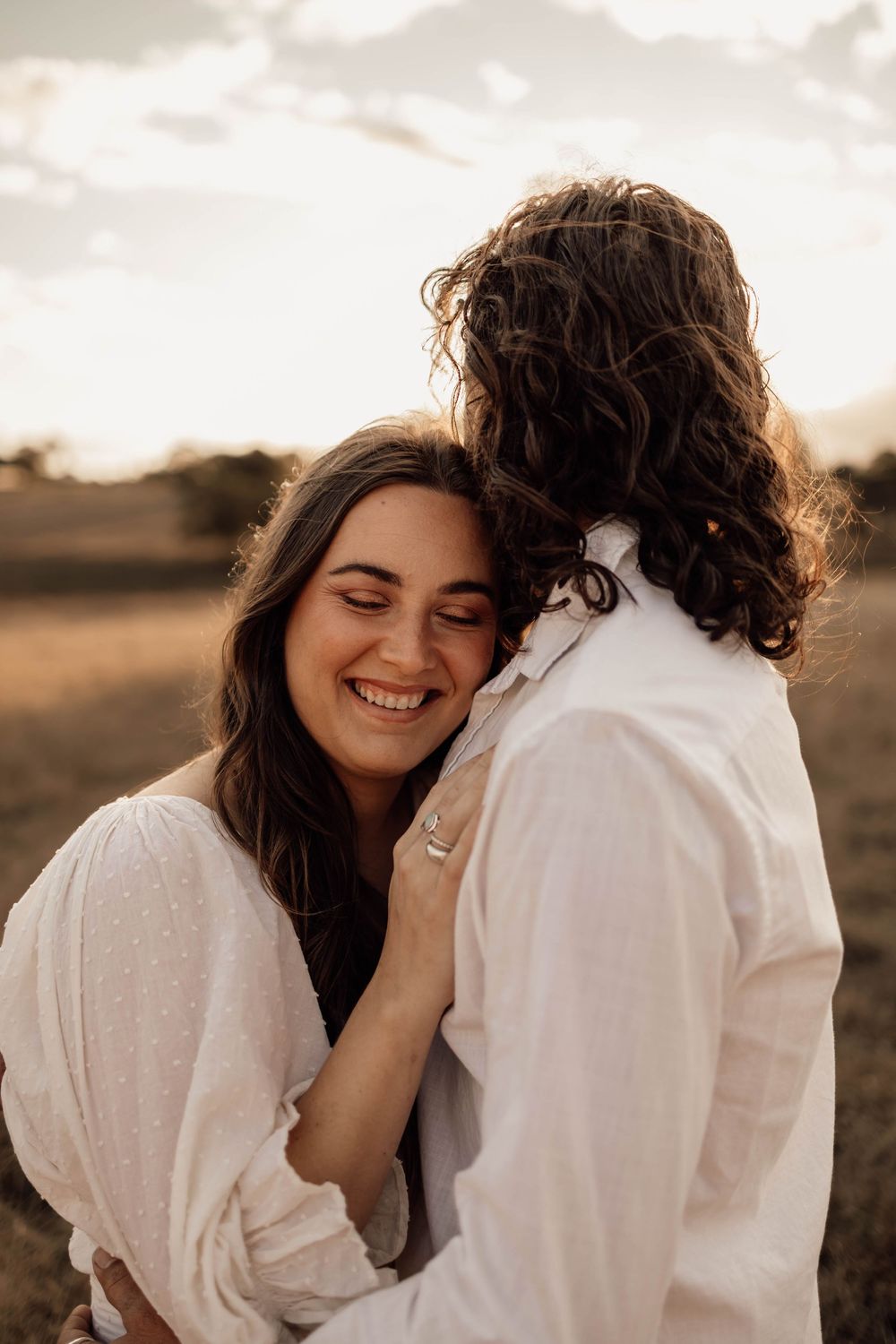 Romantic couple in white clothing embraces while sharing a tender moment in a golden sunset field.
