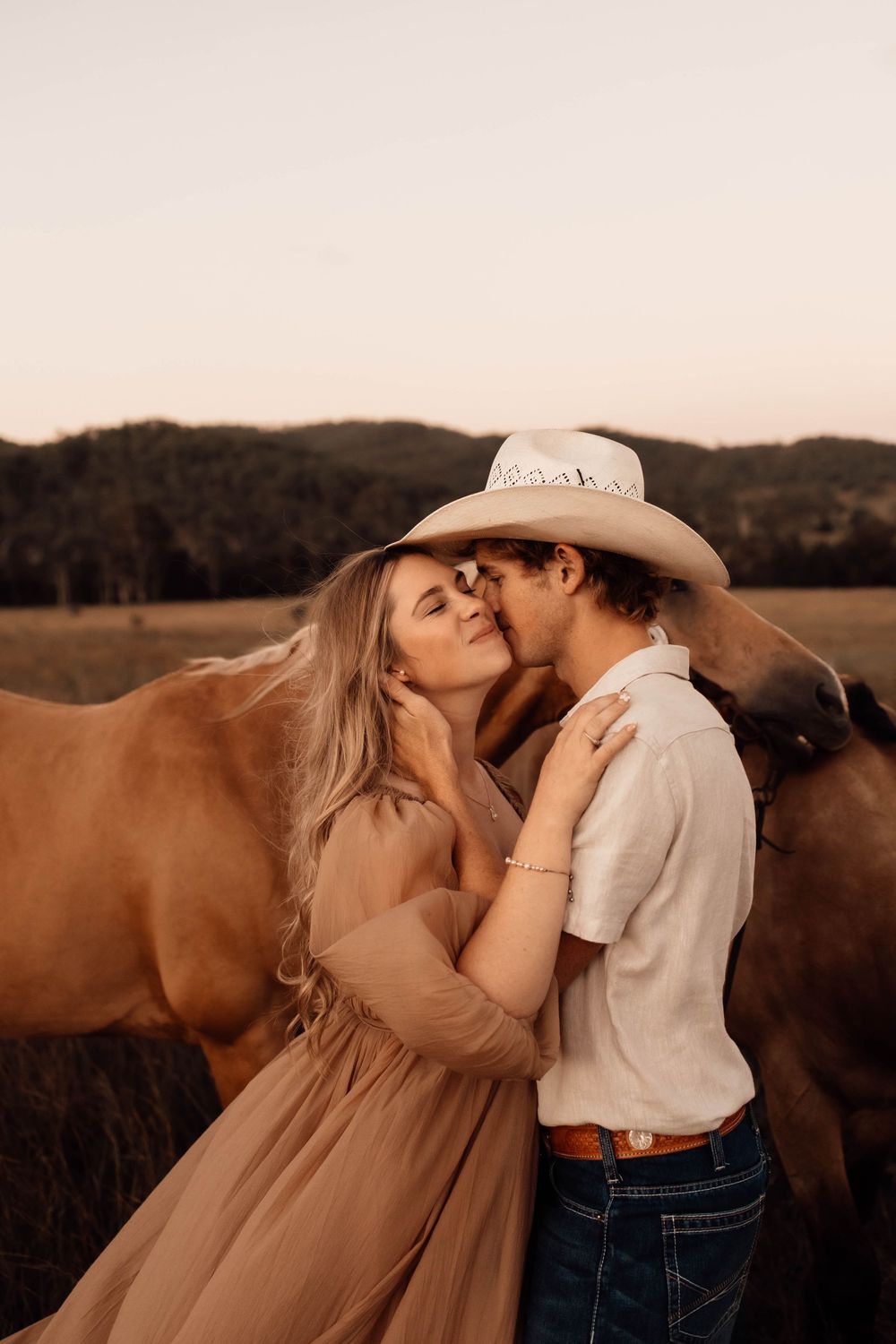 A romantic moment between a couple in a field with horses grazing in the background during sunset.