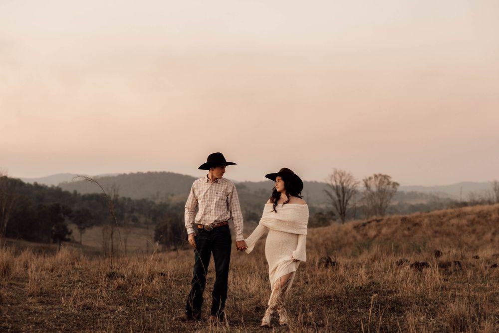 A couple in cowboy hats share an intimate moment in a golden-hour field with rolling hills behind them.