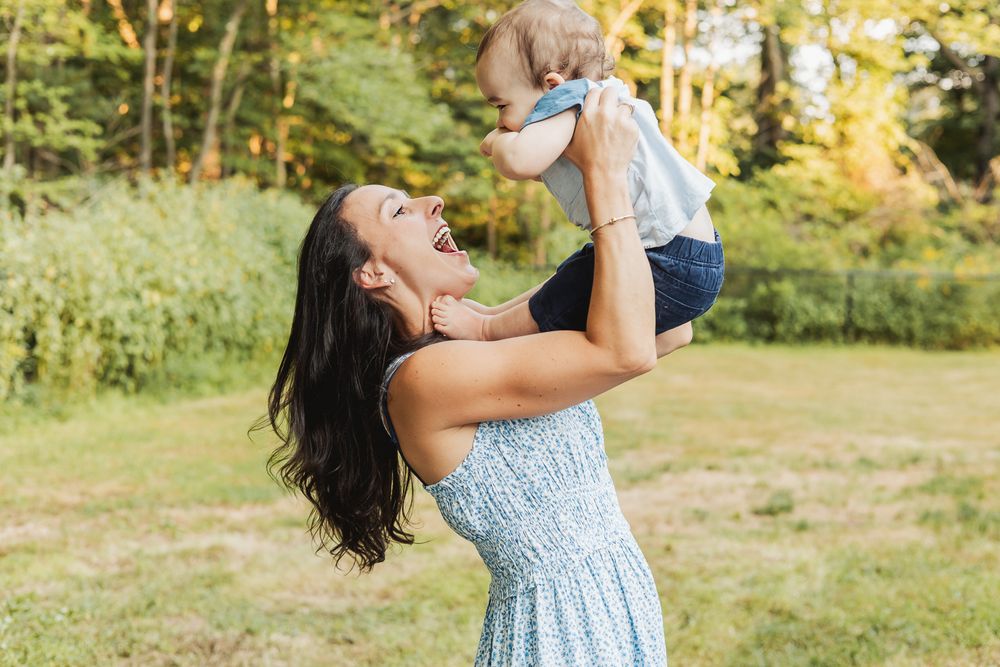 A woman in a blue floral dress lifts a small child in the air while playing outdoors on a sunny summer day.