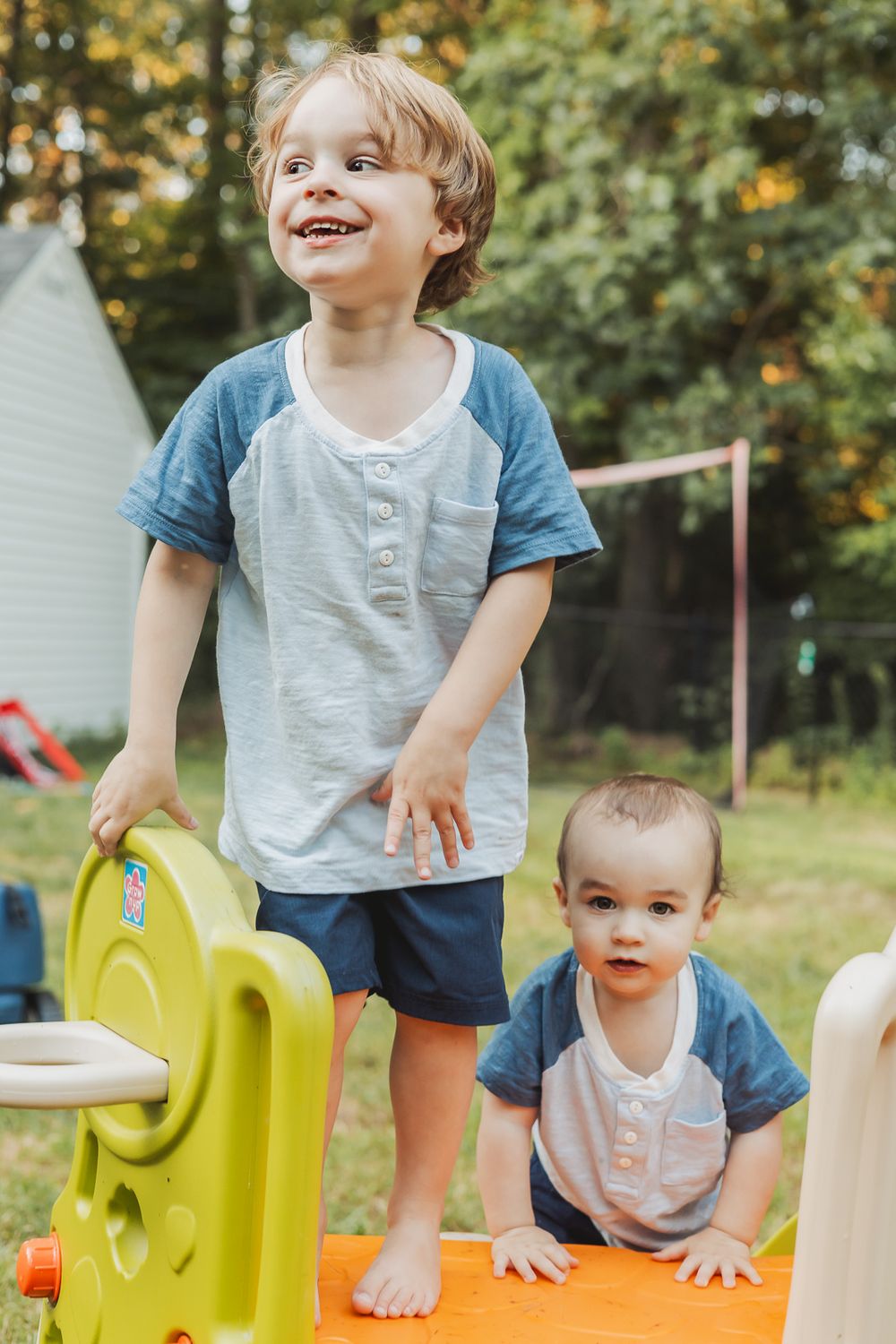 Two children playing together on a yellow plastic playground structure in a backyard.