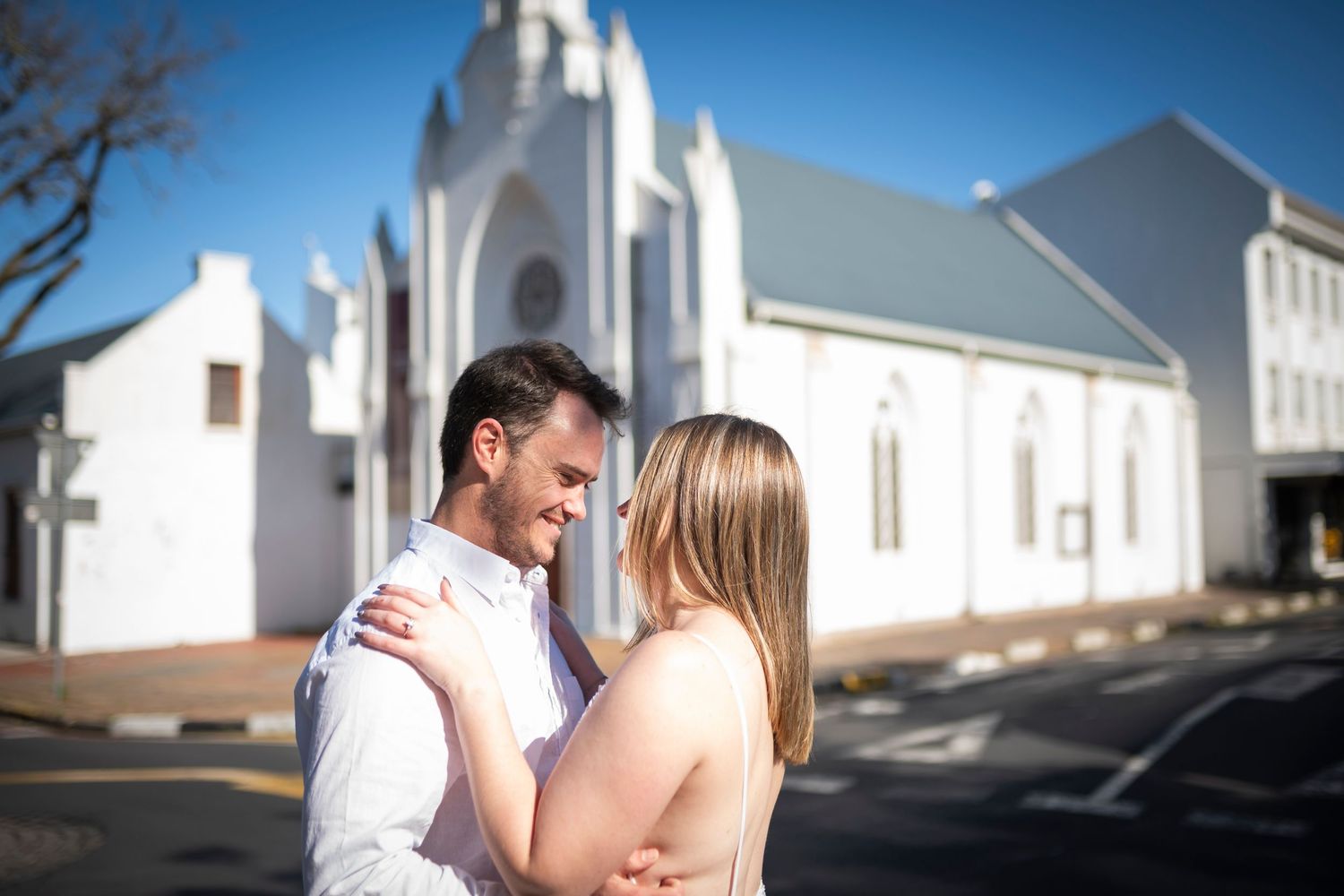 A couple shares a romantic moment outside a white church building on a sunny day.