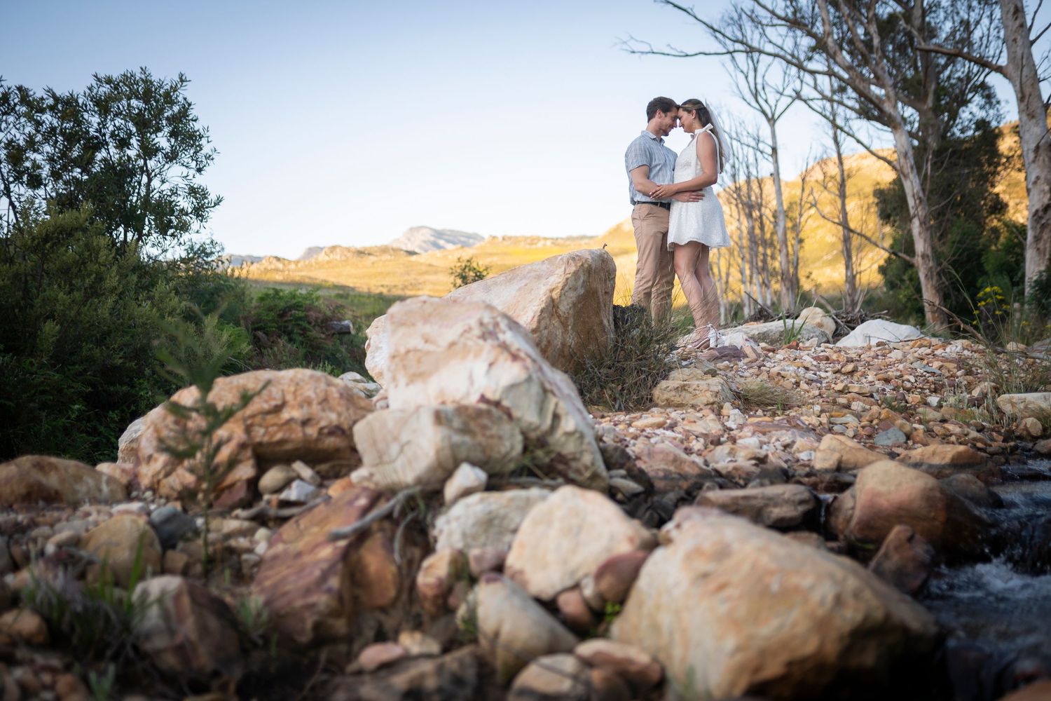 A couple stands on rocky terrain overlooking a scenic desert landscape during golden hour.