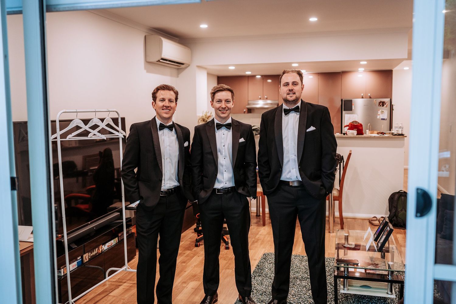 Three groomsmen in black tuxedos and bowties pose together in a formal setting.