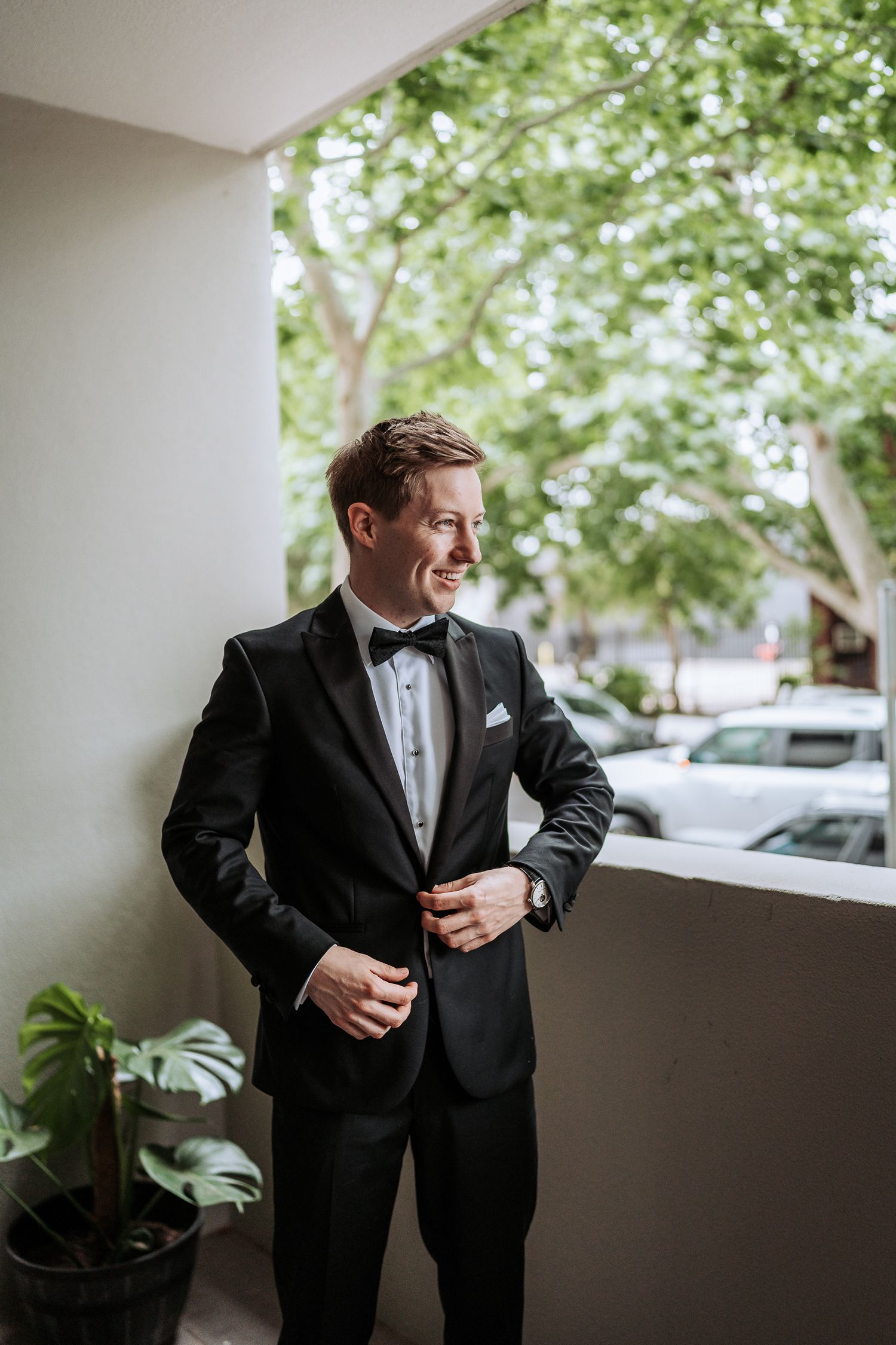 A groom in a classic black tuxedo adjusts his jacket while standing in front of a window with greenery visible outside.