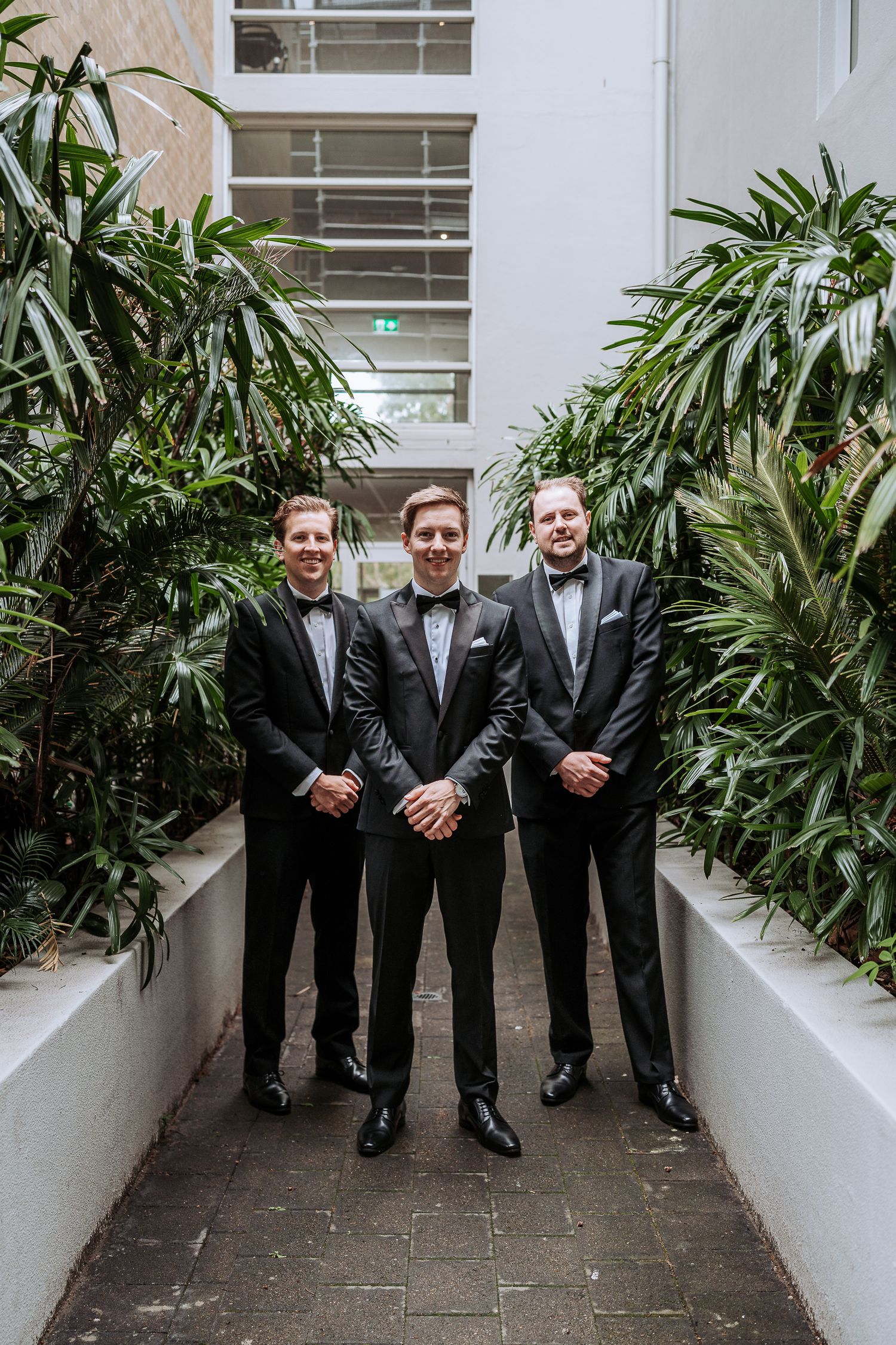 Groomsmen in black tuxedos pose together in a modern hallway with tropical plants along white walls.