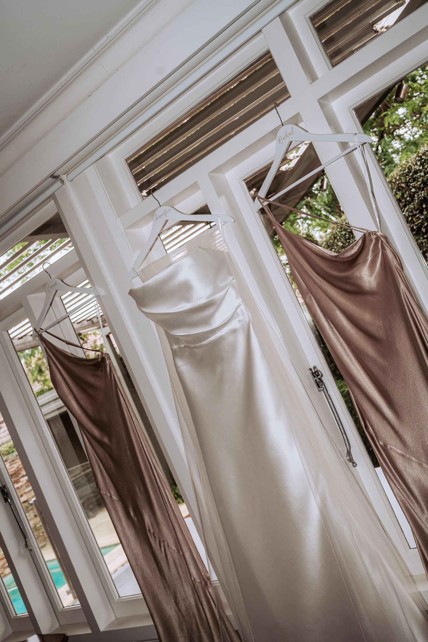Elegant silk dresses in neutral tones hanging in a row against white window frames with natural lighting.