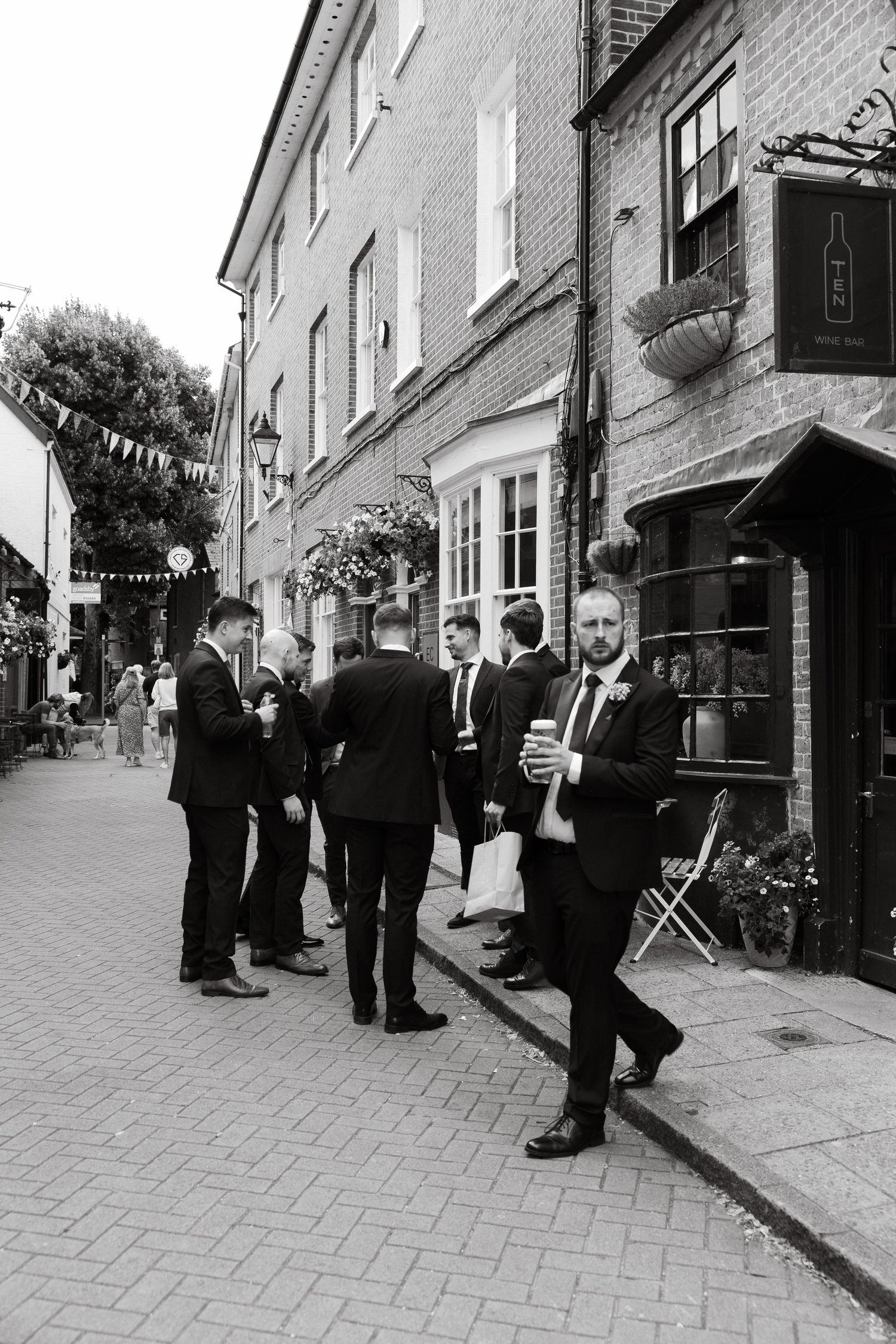 Group of formally dressed people gathering outside brick buildings on a cobblestone street.