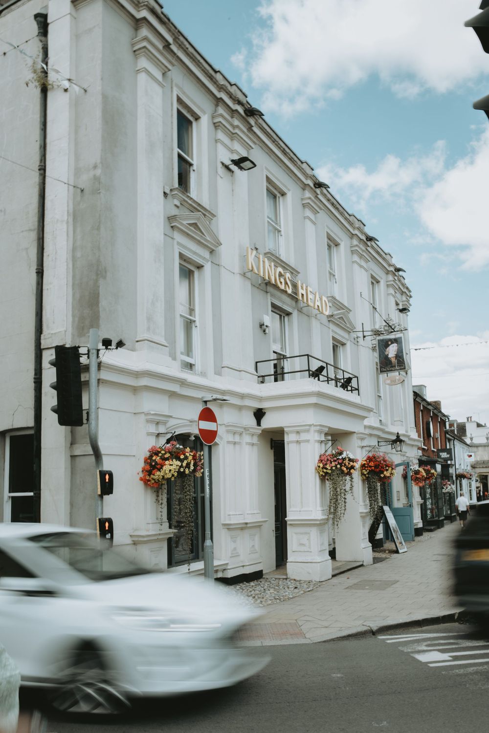 White historic hotel building with hanging flower baskets and a car passing by on a sunny day.