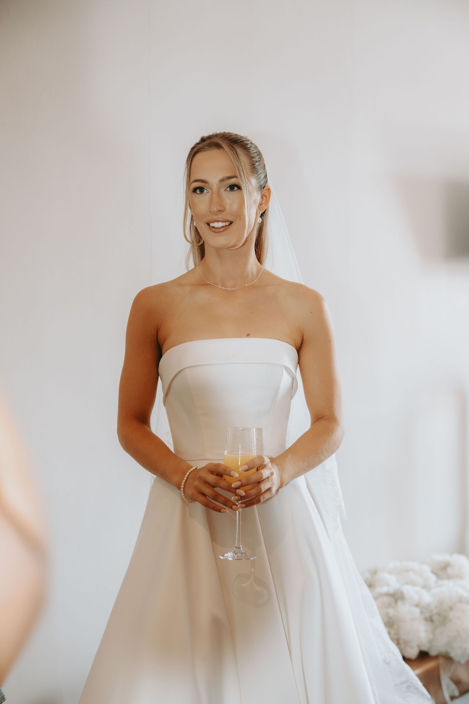 A person wearing a strapless white wedding gown holds a champagne glass while getting ready.