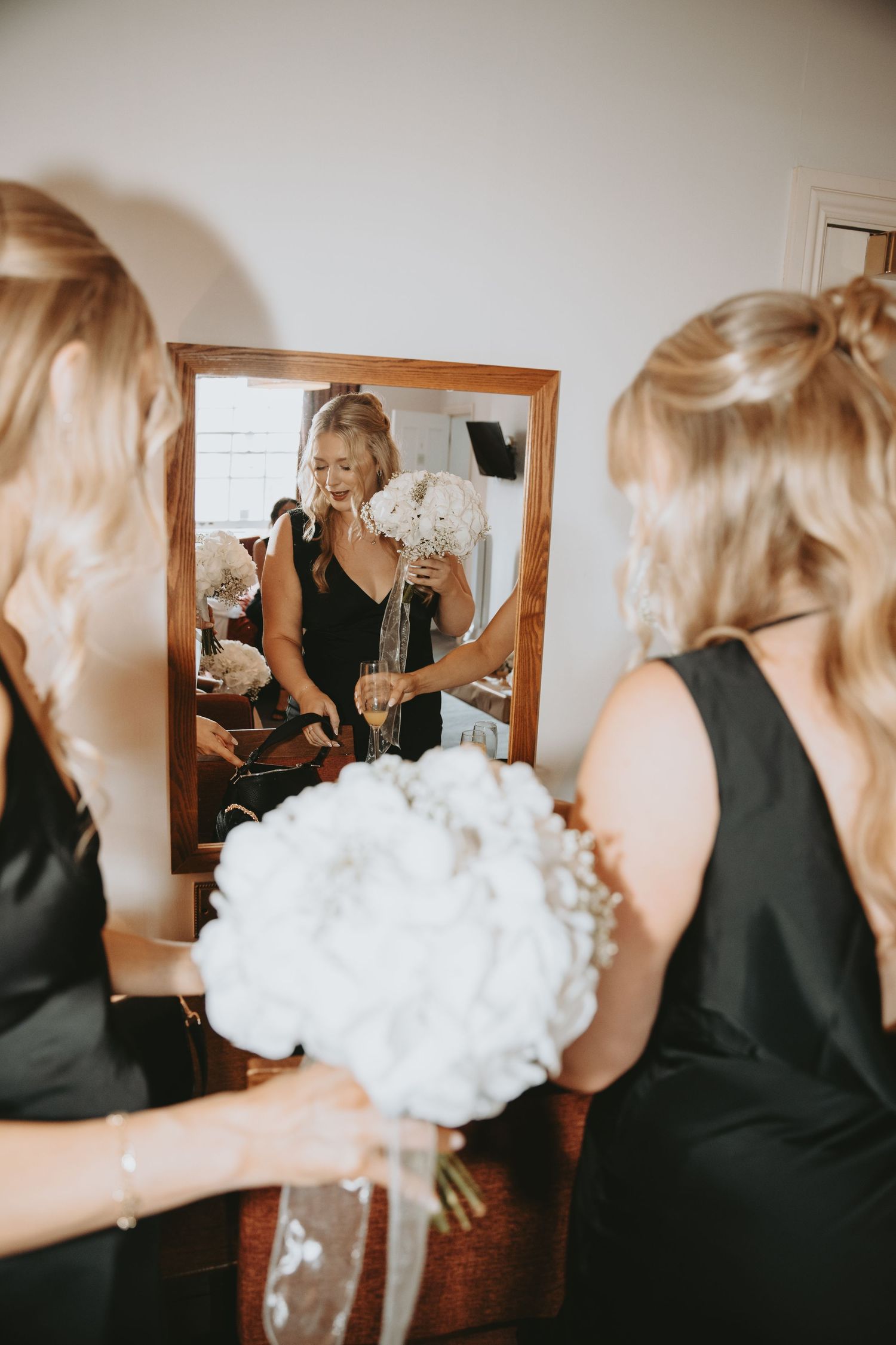 Wedding party members in black dresses hold white floral bouquets while getting ready.