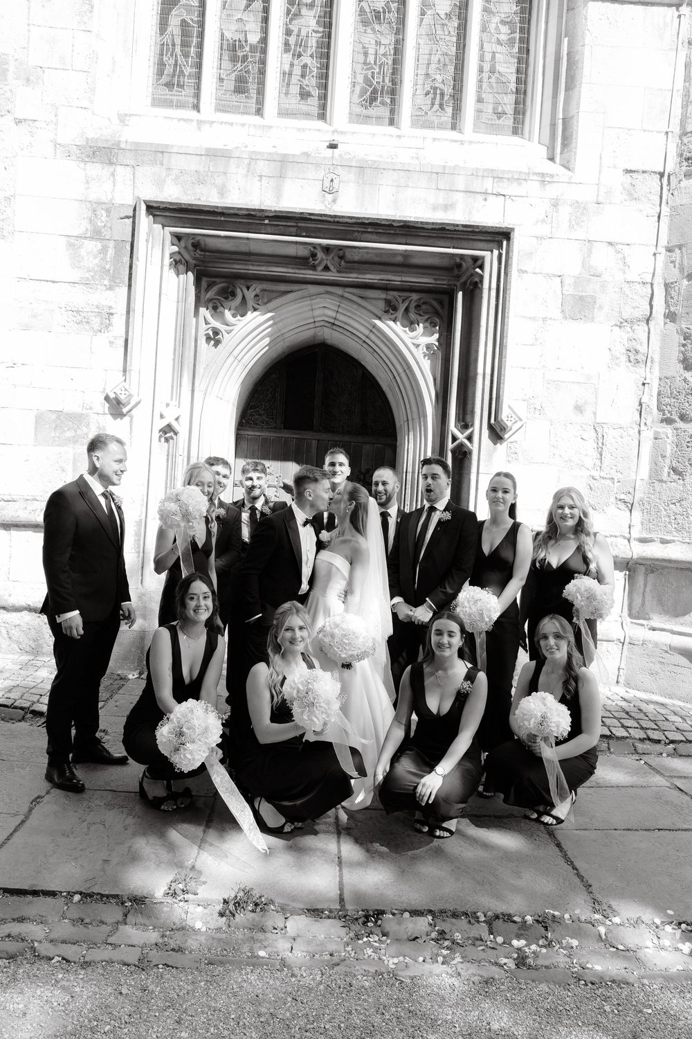 Wedding party posing together outside historic stone church entrance.