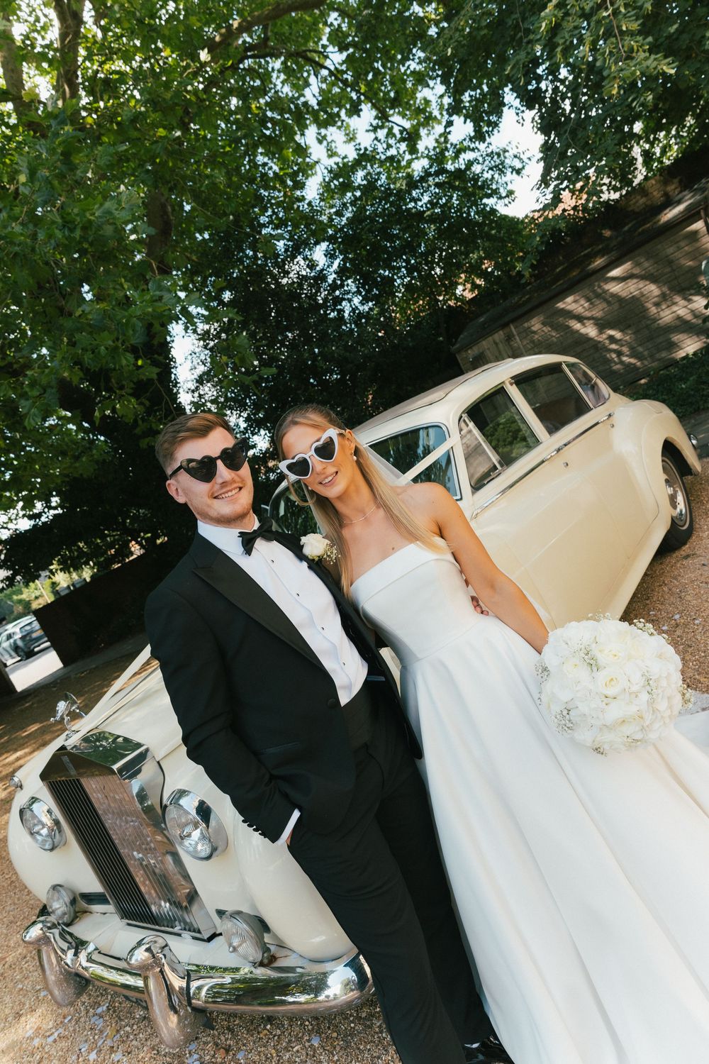 Wedding couple posing with classic white vintage car under green trees.