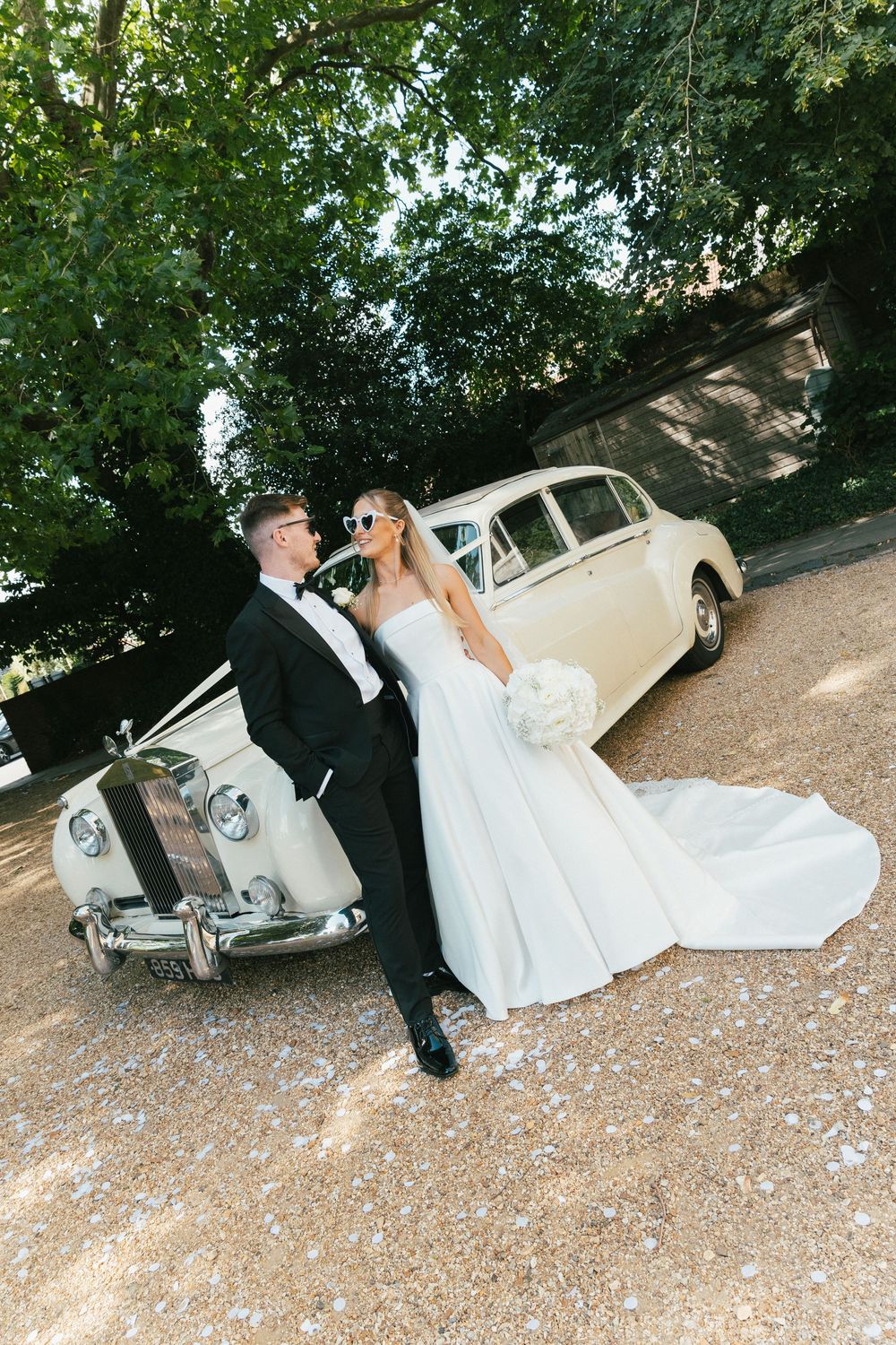 Newlyweds in sunglasses relaxing by vintage white car on gravel path.