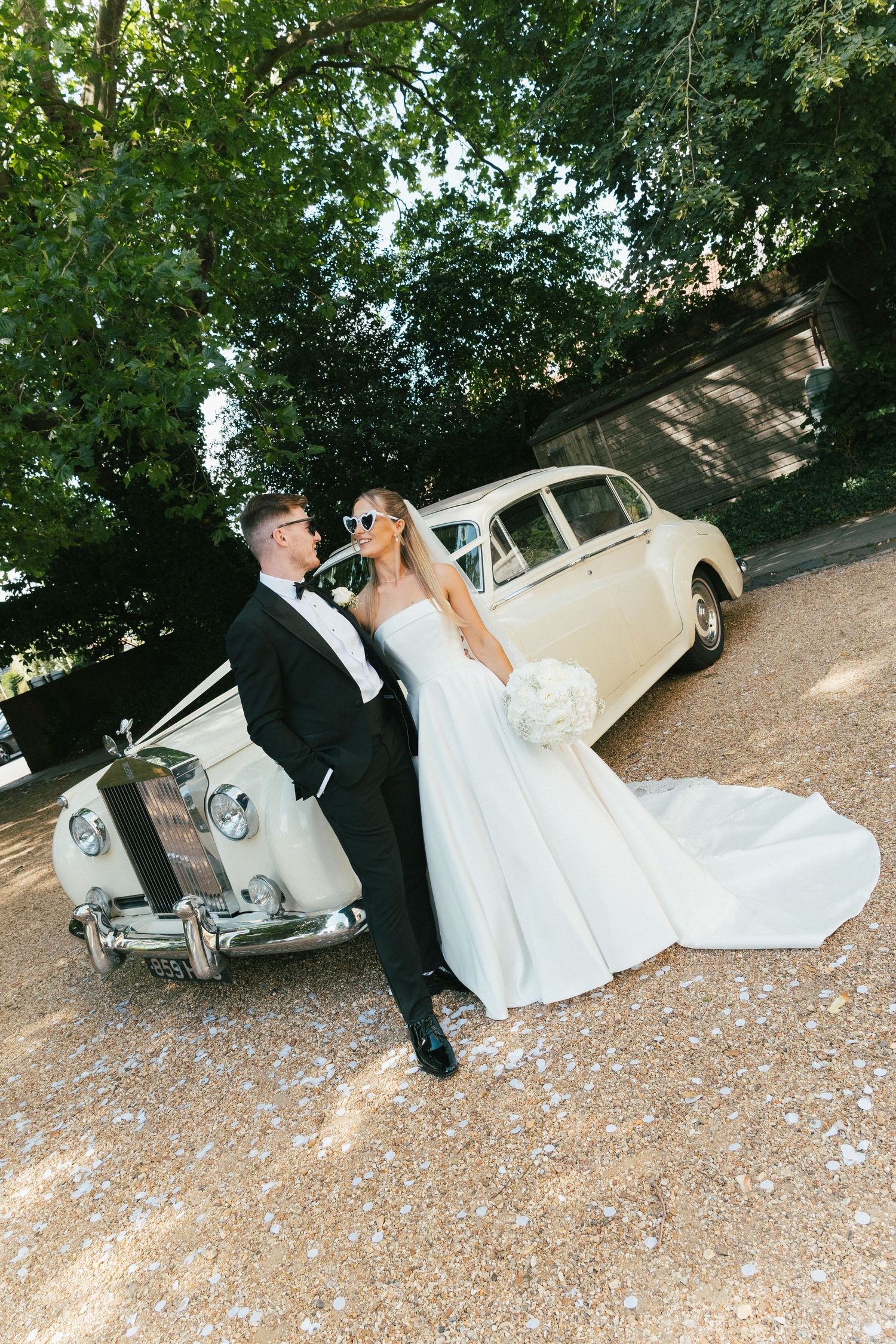 Newlyweds in sunglasses relaxing by vintage white car on gravel path.