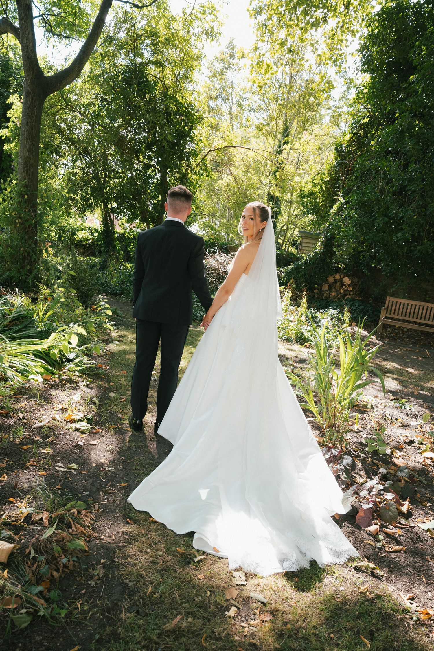 A wedding couple walks through a lush garden setting with dappled sunlight.