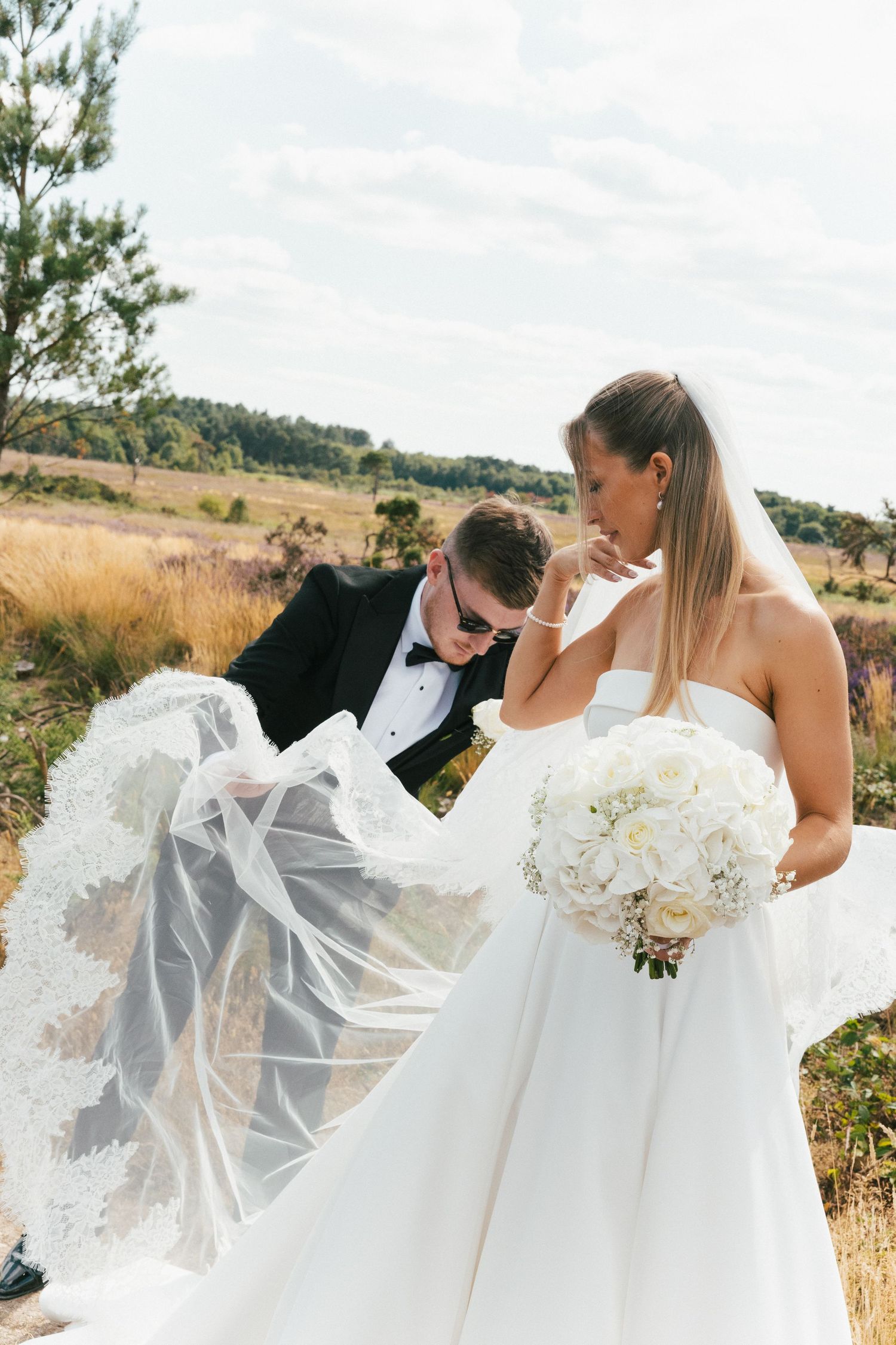 Wedding couple embracing in golden field with flowing veil catching the breeze.