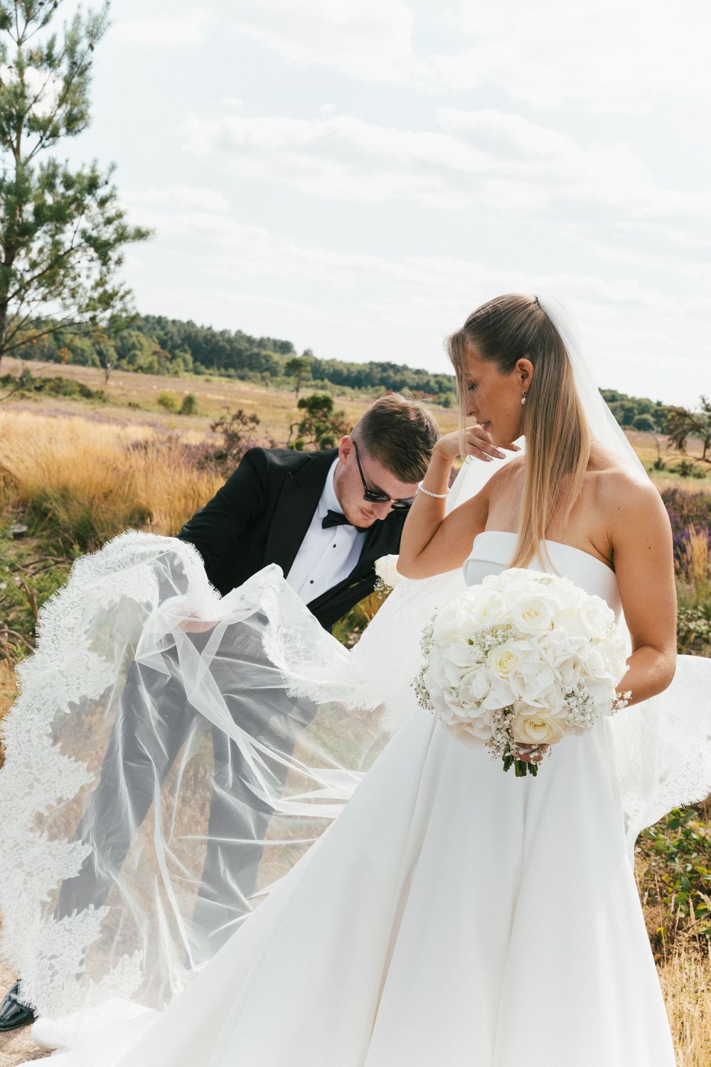 Wedding couple embracing in golden field with flowing veil catching the breeze.