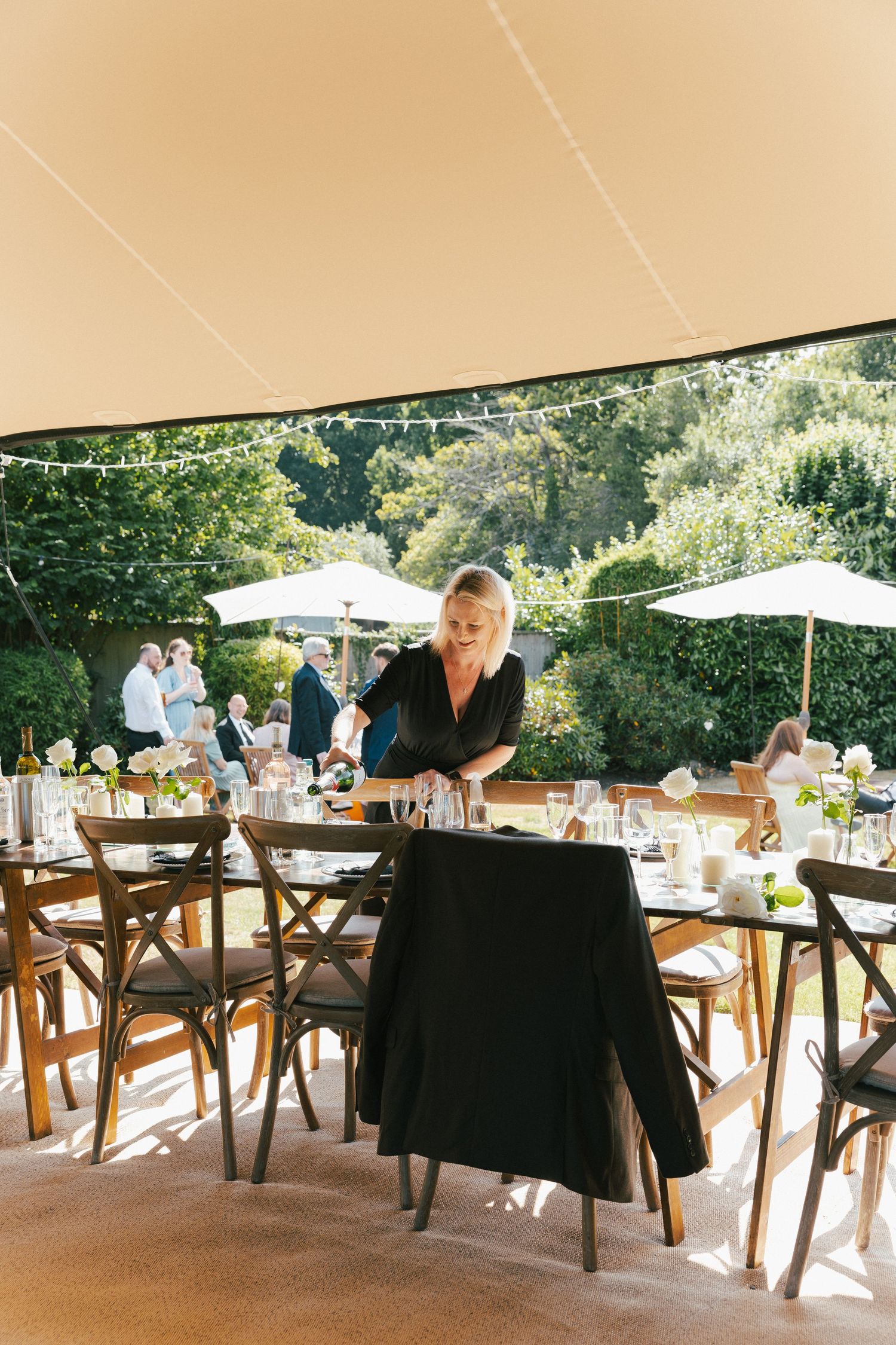 An outdoor wedding reception setup with wooden cross-back chairs and white tablecloths.