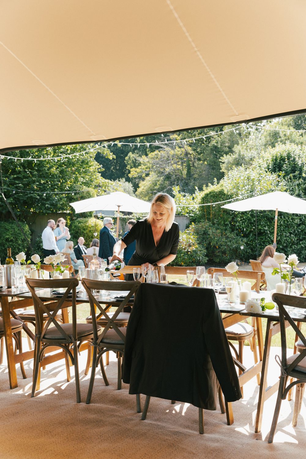 An outdoor wedding reception setup with wooden cross-back chairs and white tablecloths.