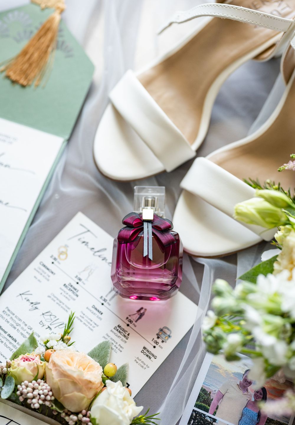 Wedding details including pink perfume bottle, white flowers, and invitation cards arranged on a light gray surface.