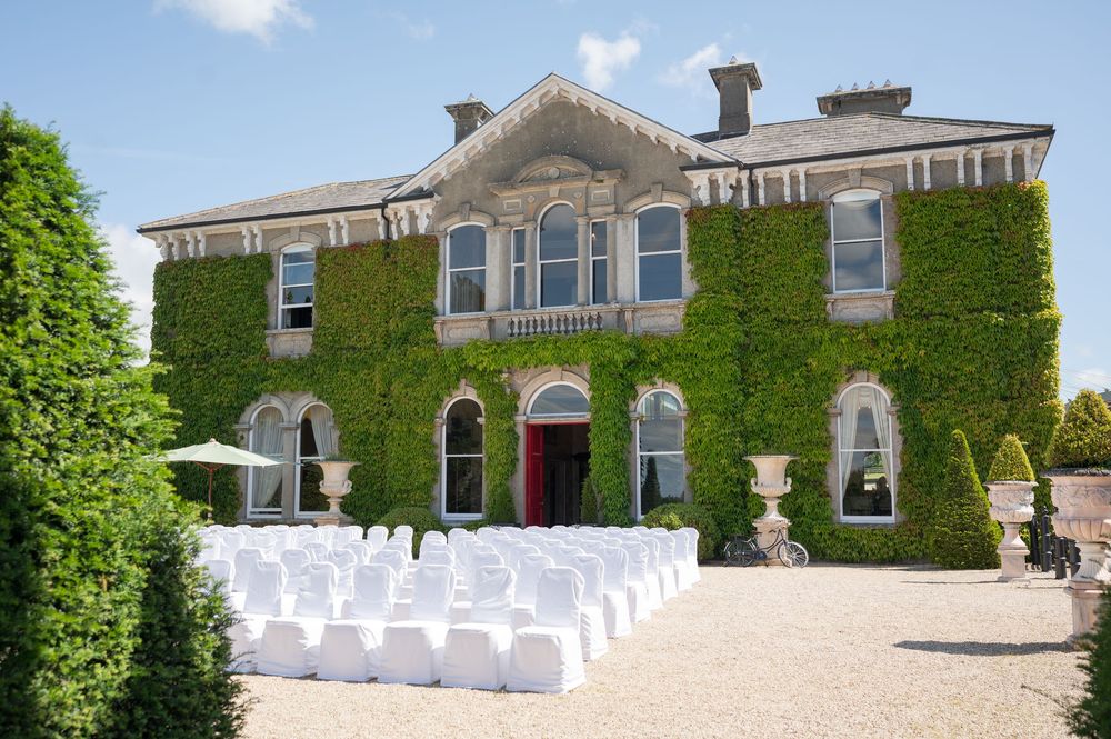 Elegant ivy-covered mansion with white chairs set up for an outdoor wedding ceremony on a gravel courtyard.