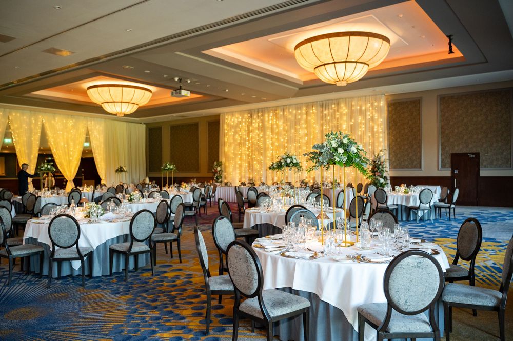Elegant ballroom setup with round tables, white linens, and ornate chairs under warm lighting and crystal chandeliers.