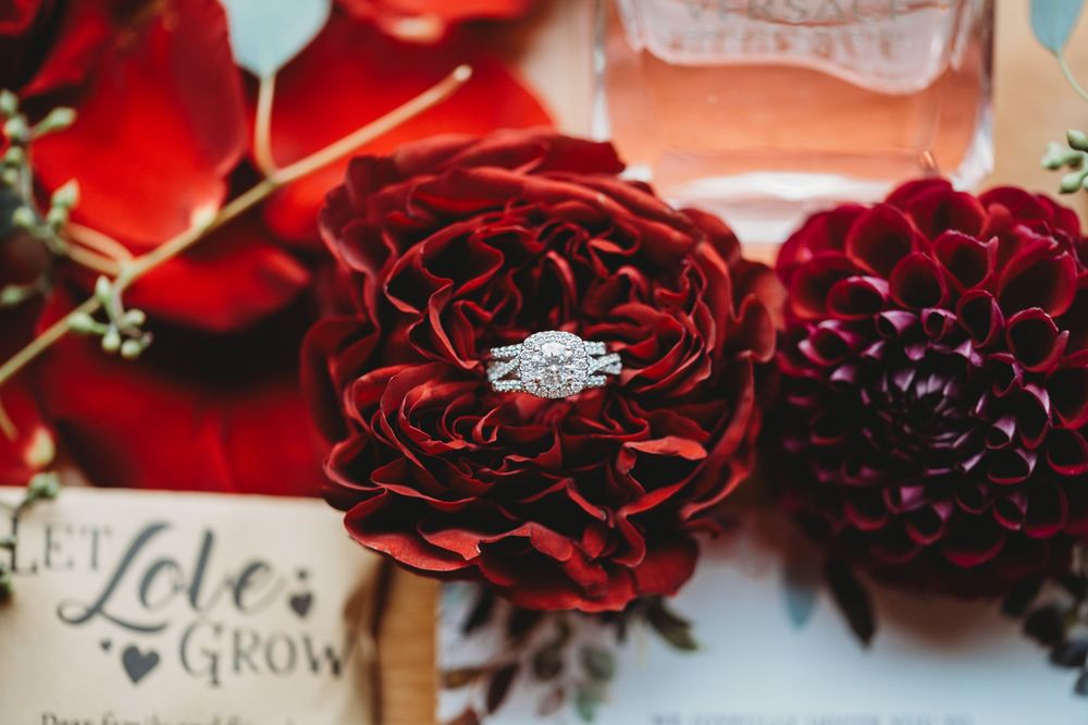 A sparkling diamond engagement ring nestled in deep red dahlia flowers with vintage style decor elements.