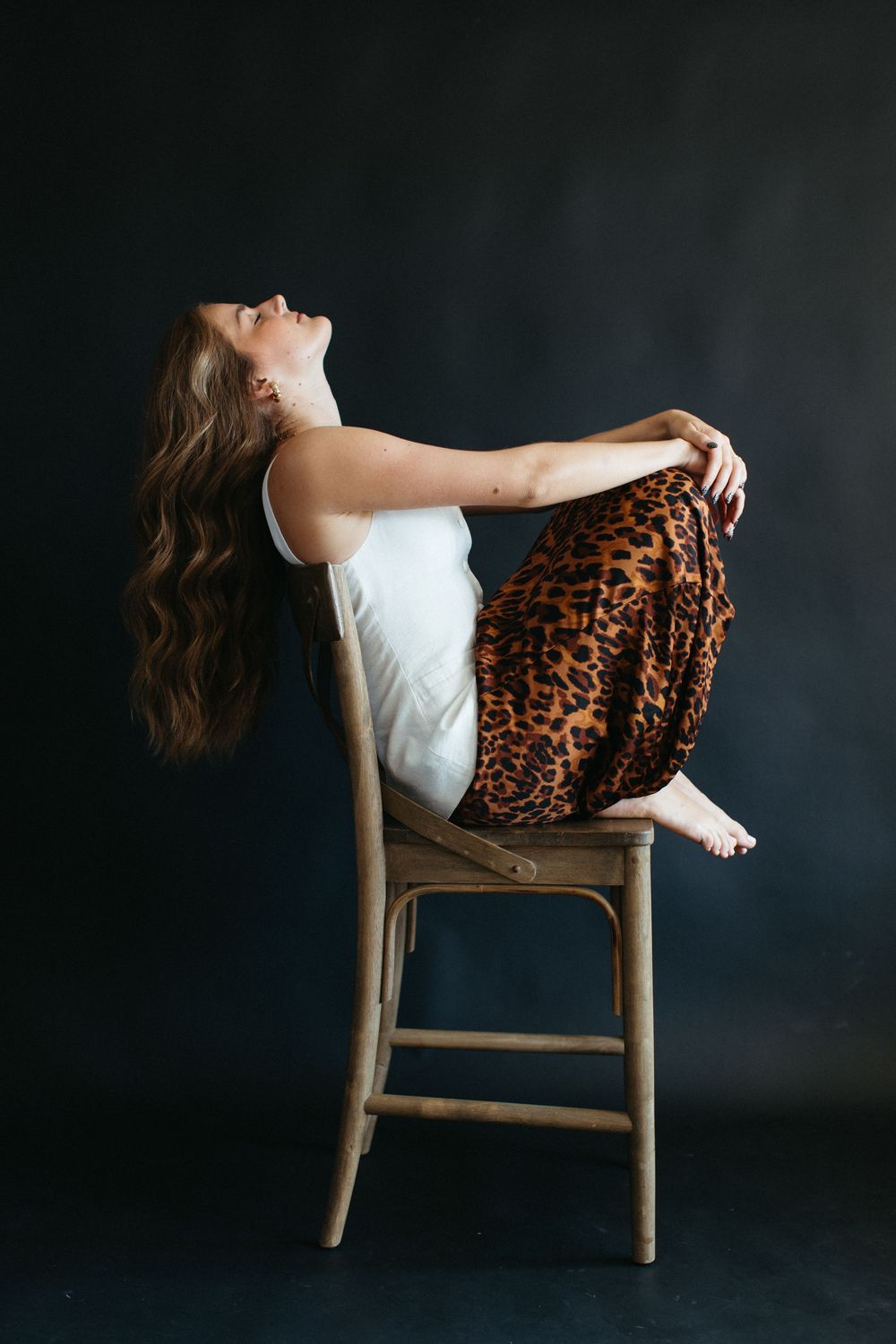 Artistic portrait of someone in white top and leopard print skirt leaning back on wooden chair against dark background.
