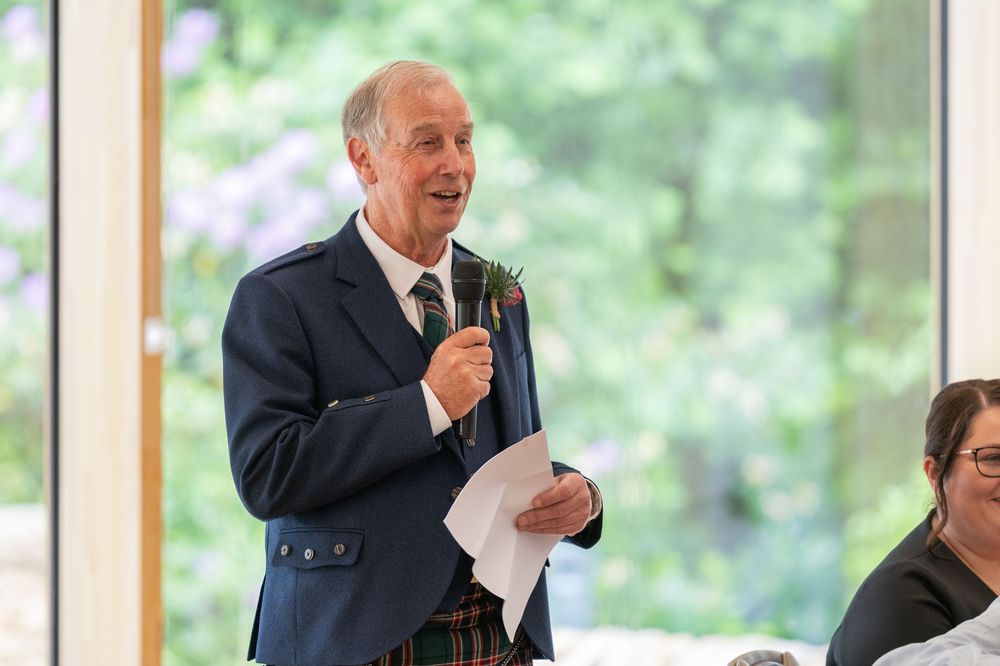 A person speaks into a microphone while addressing guests at a formal event in a bright, window-lined venue.