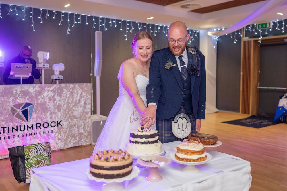 Couple stands behind a table with three tiered cakes at their wedding reception with twinkle lights in background.