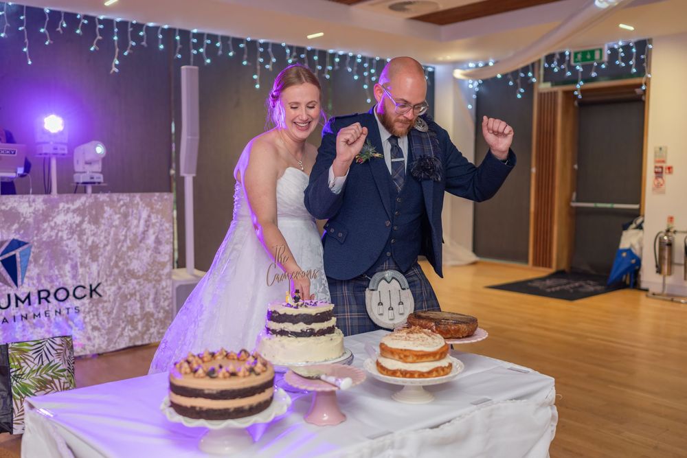 Wedding couple celebrates enthusiastically while standing behind their display of three-tiered cakes at the reception.