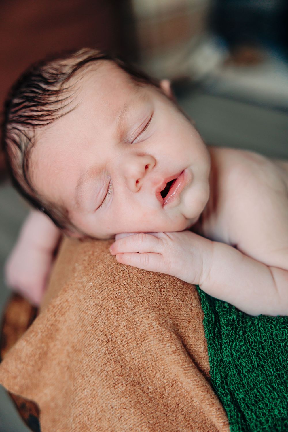 A sleeping newborn baby rests peacefully on brown and green textured blankets.