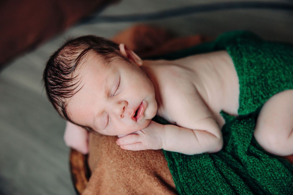 Sleeping newborn wrapped in emerald green fabric rests peacefully on brown blanket during professional photoshoot.