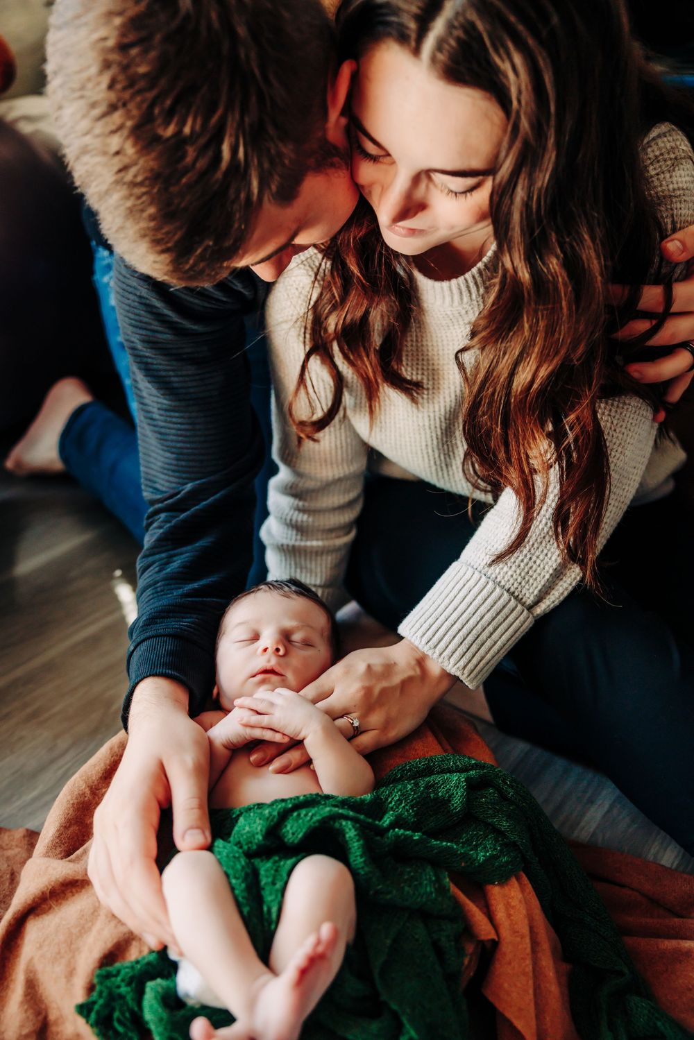 Parents tenderly interact with their newborn baby wrapped in an emerald green blanket on hardwood floor.