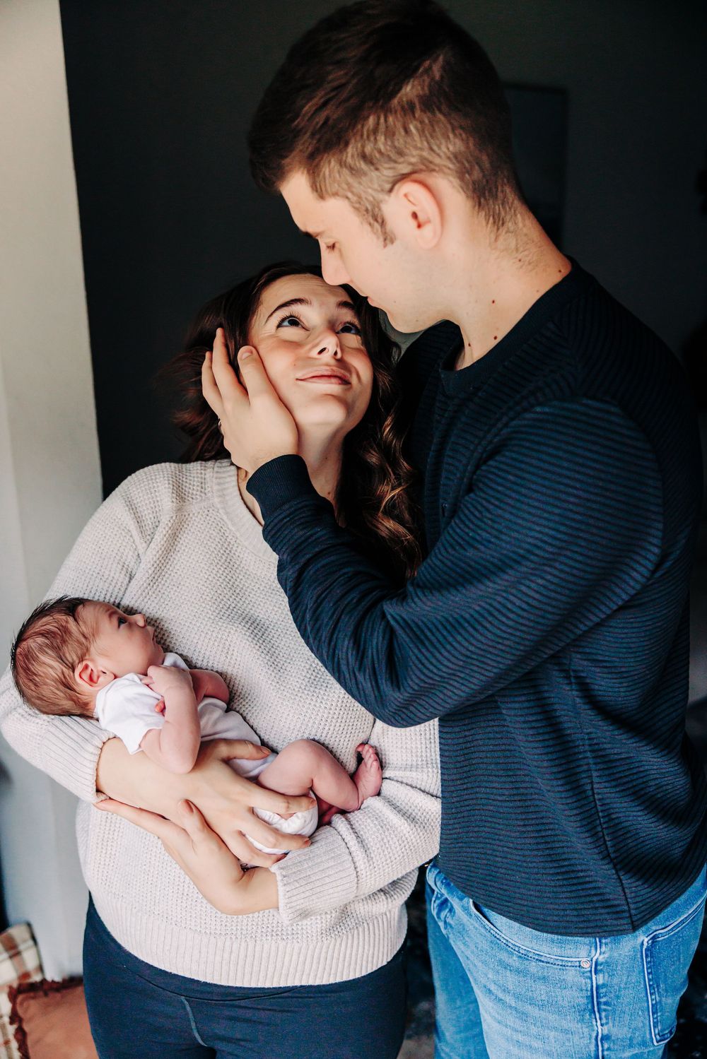 Parents in blue jeans and sweaters tenderly bonding with their newborn baby against dark background.