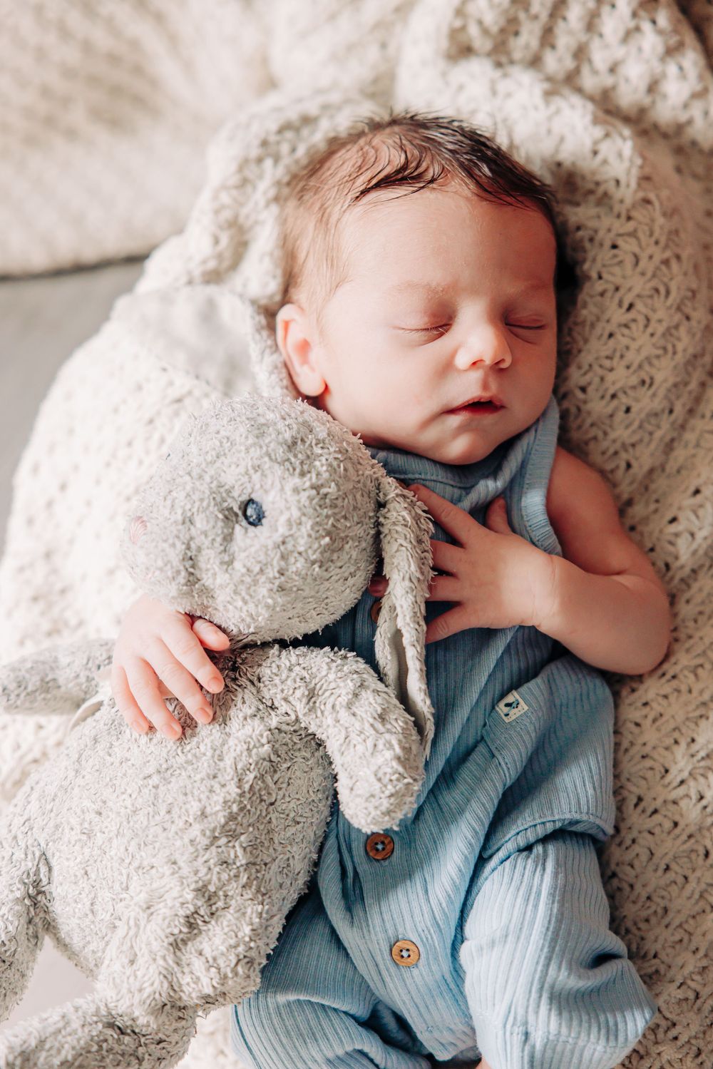 A sleeping baby in blue clothing snuggles with a plush bunny toy on soft white bedding.