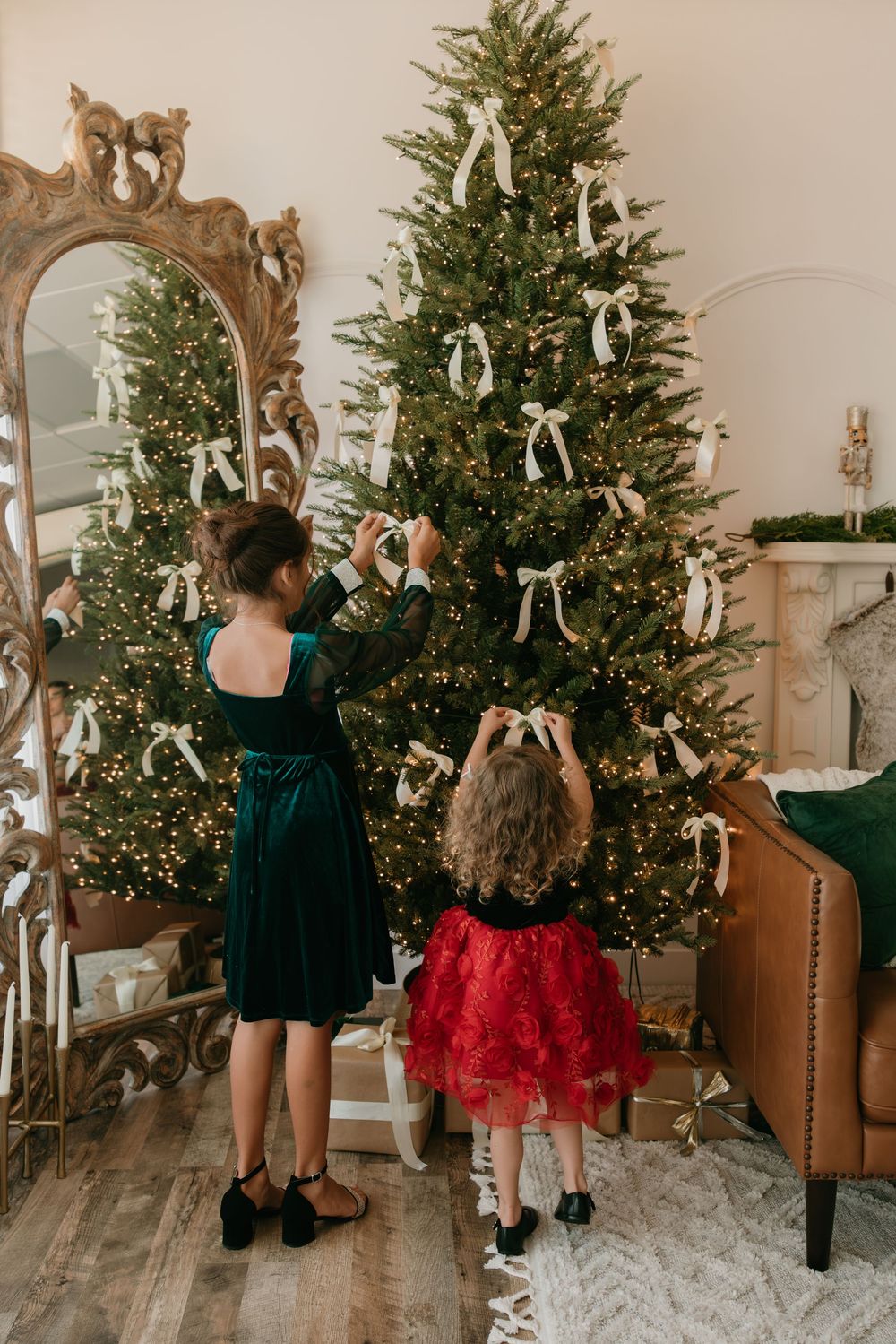 Two children in holiday outfits look at a decorated Christmas tree with white bows and twinkling lights.