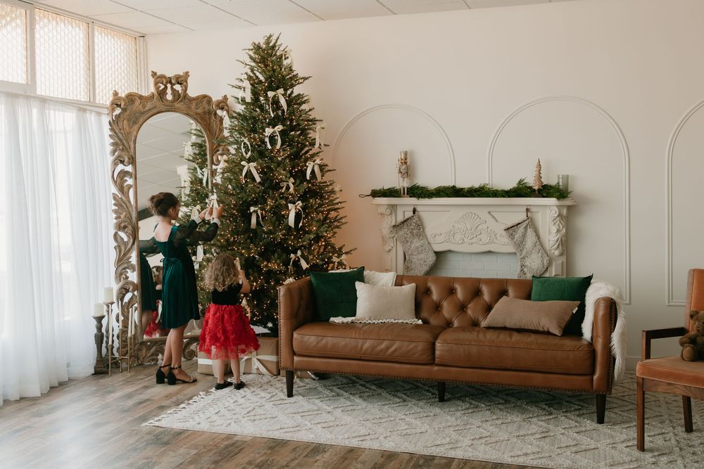 A bright modern living room decorated for Christmas with multiple trees, white walls, and brown leather furniture.