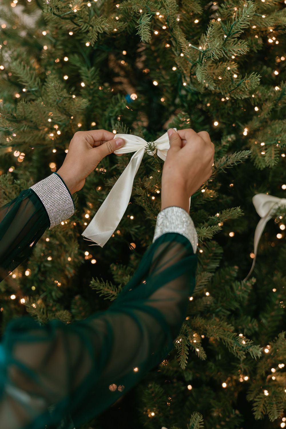 Hands tying a white ribbon bow on a Christmas tree with twinkling lights in the background.