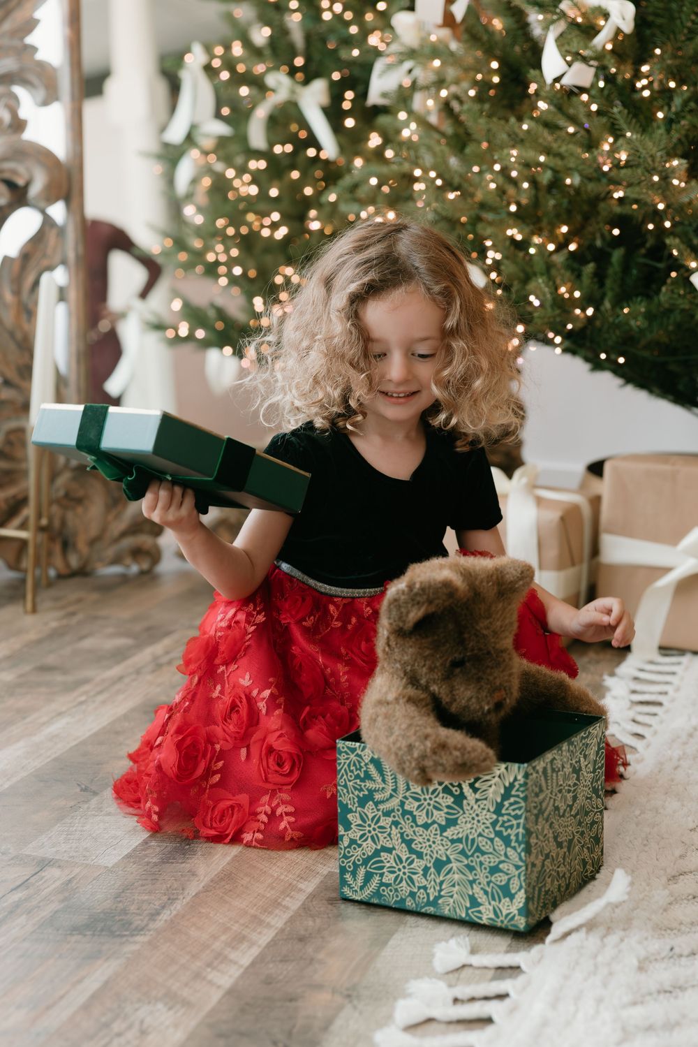 A child in a red tulle skirt opens Christmas presents near a decorated tree with a teddy bear and gift boxes.