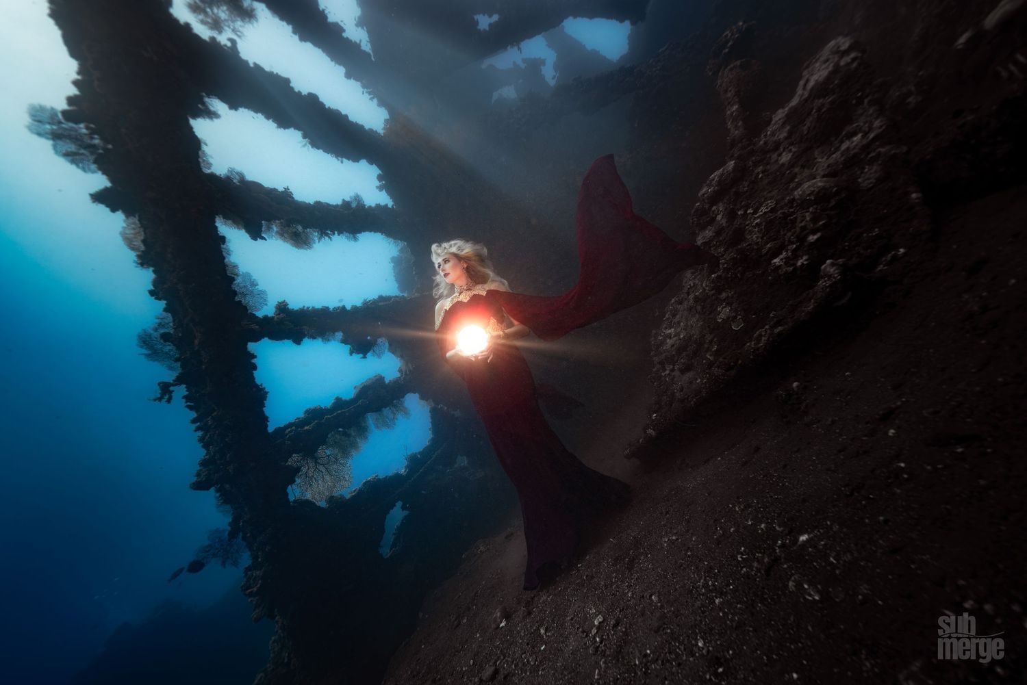 Underwater diver swims beneath a dark blue surface with dramatic sunlight streaming through the water.