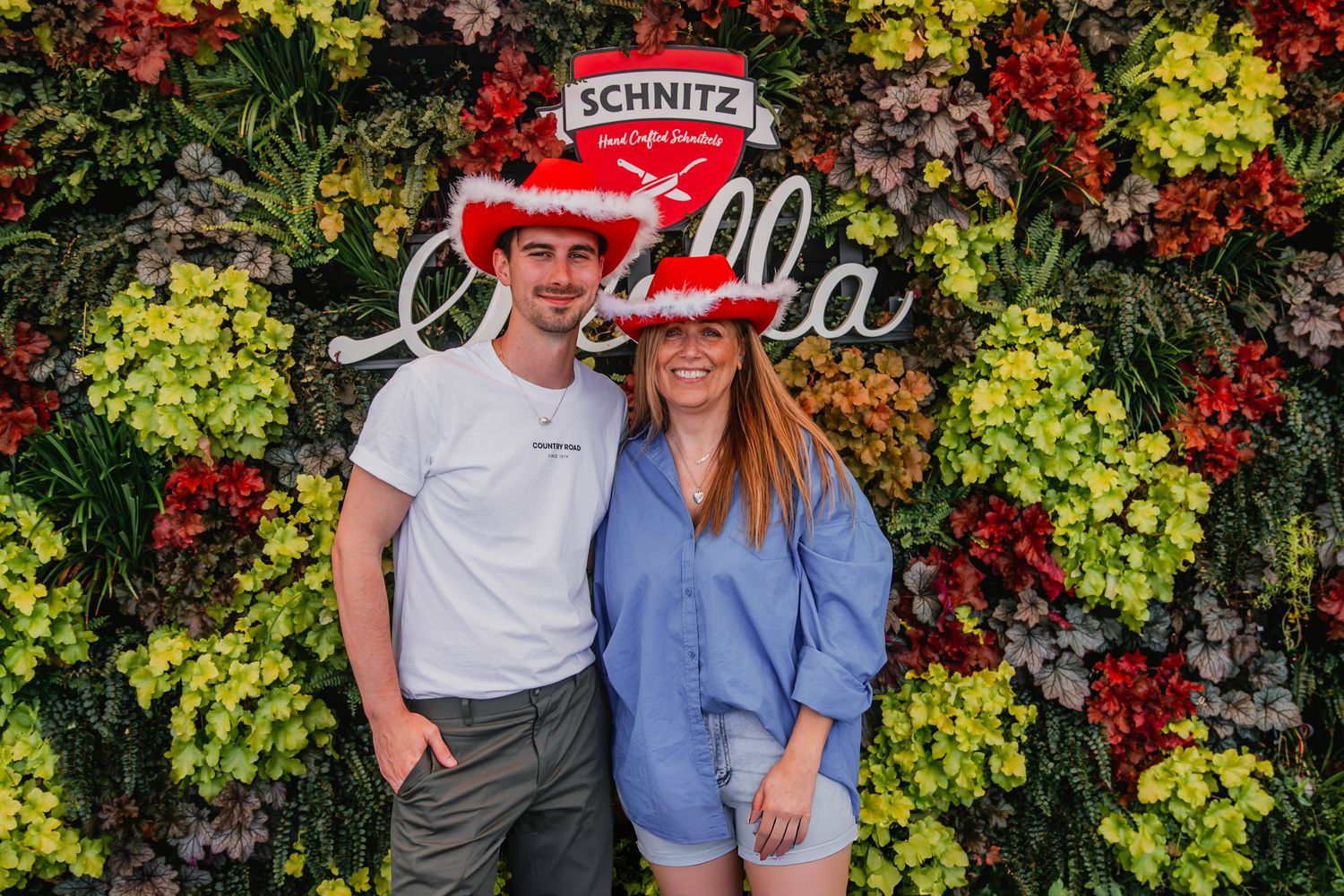 Two people pose together against a vibrant autumn foliage wall with colorful red and yellow leaves.