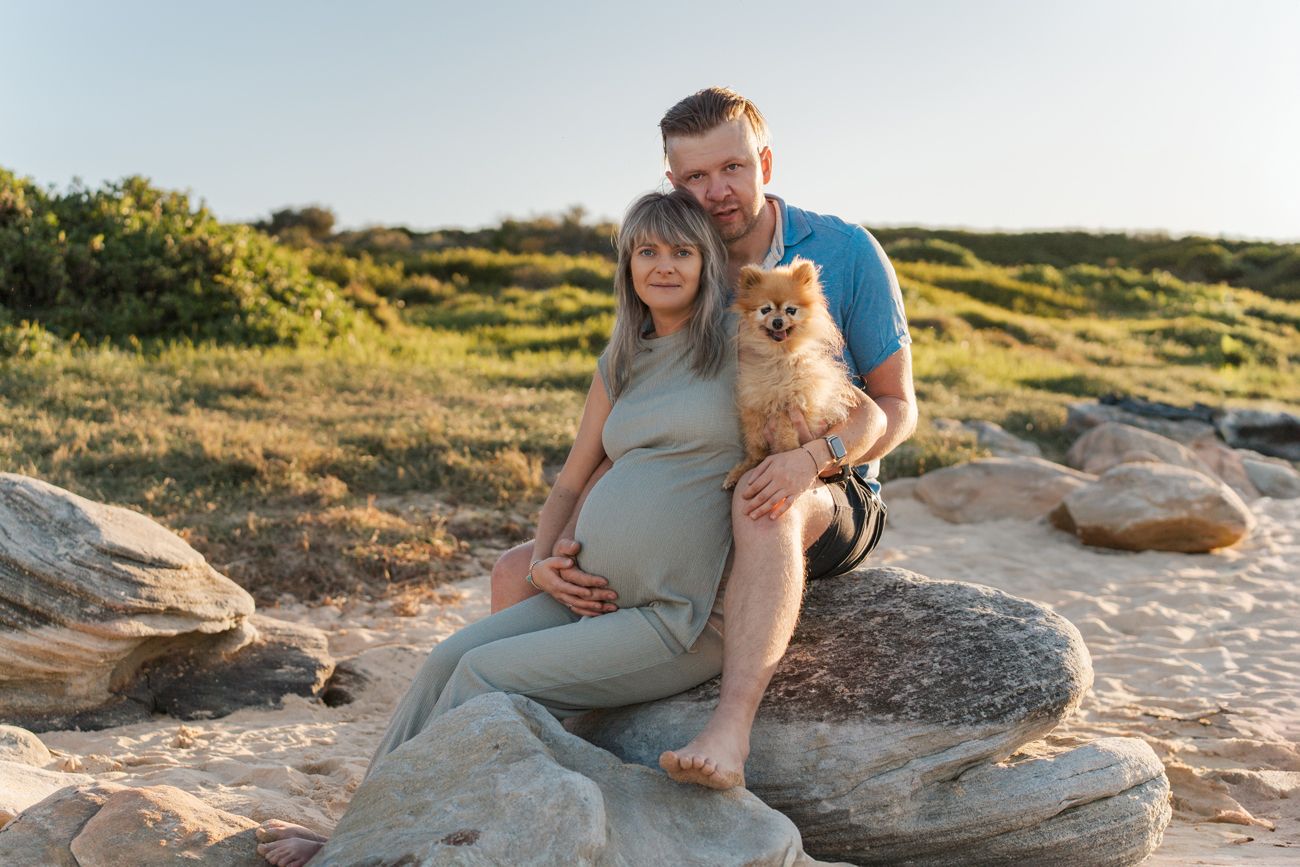 A couple sits on coastal rocks at sunset with their Pomeranian dog as they pose for a maternity photo on Maroubra Beach in Sydney Photoshoot