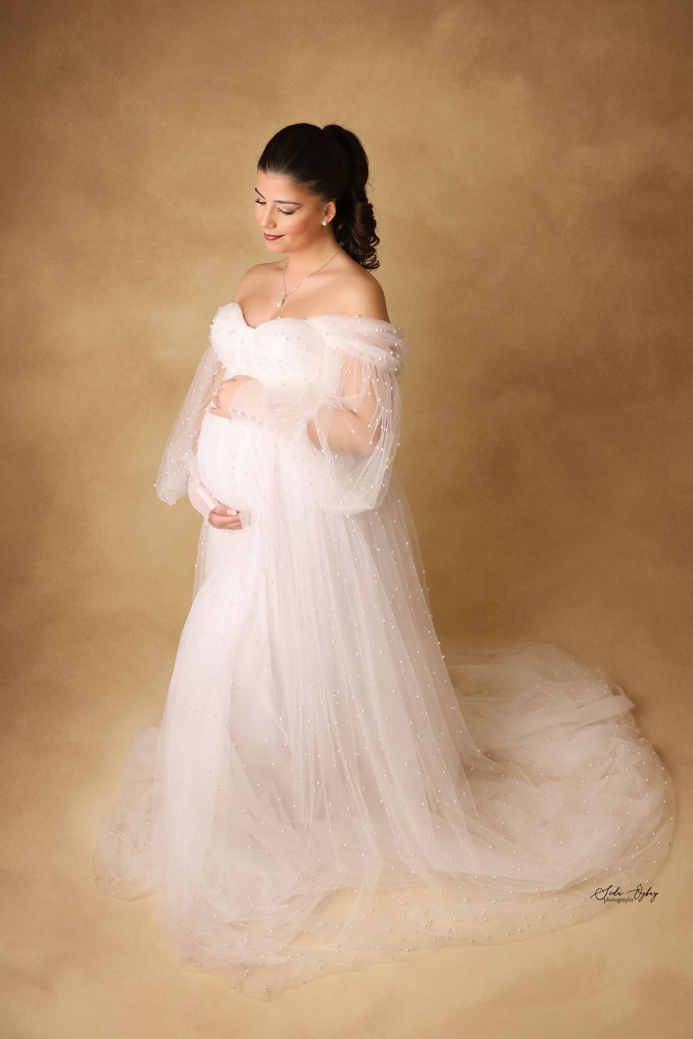 Dreamy maternity portraits in flowing white tulle gown against warm brown studio backdrop.