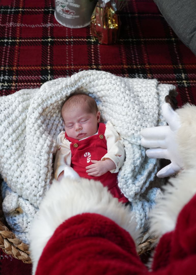 Baby in red outfit rests peacefully on white blanket next to white cat during holiday season.