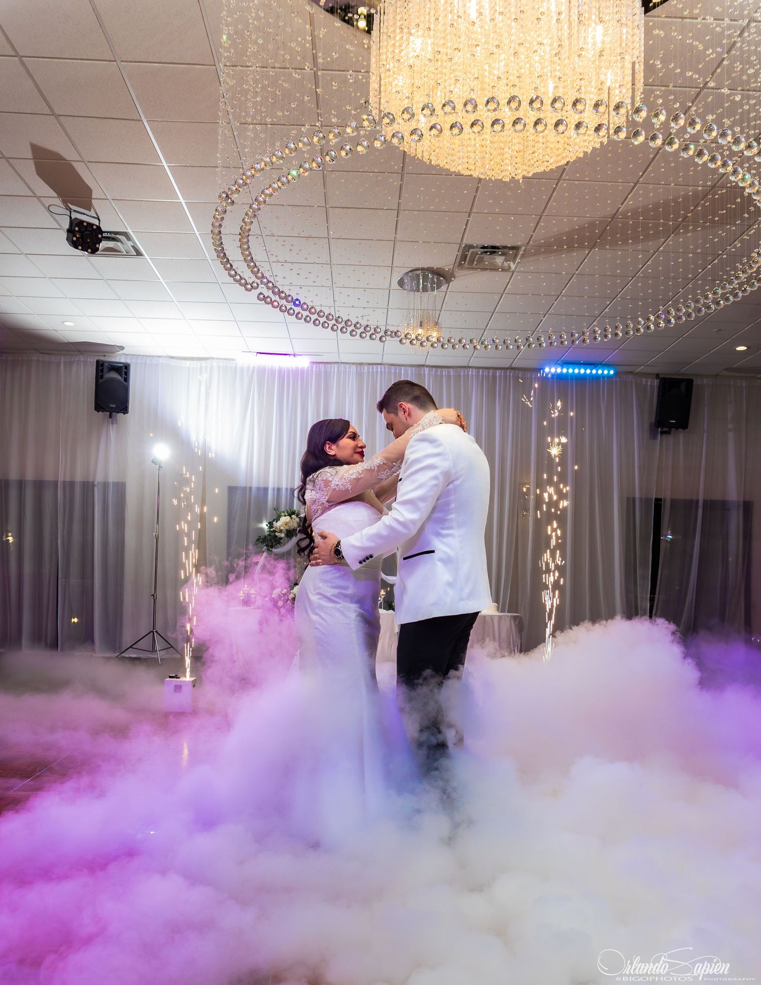 Couple dances their first dance surrounded by dramatic purple fog effects on an elegant ballroom floor with chandeliers above.