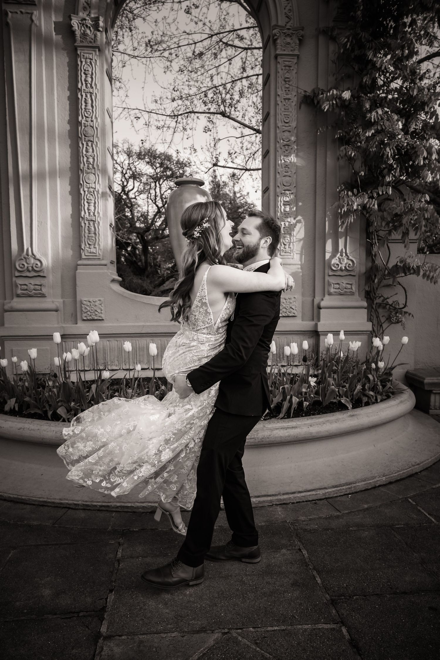 A couple in wedding attire walks along a scenic garden path surrounded by lush greenery and trees.